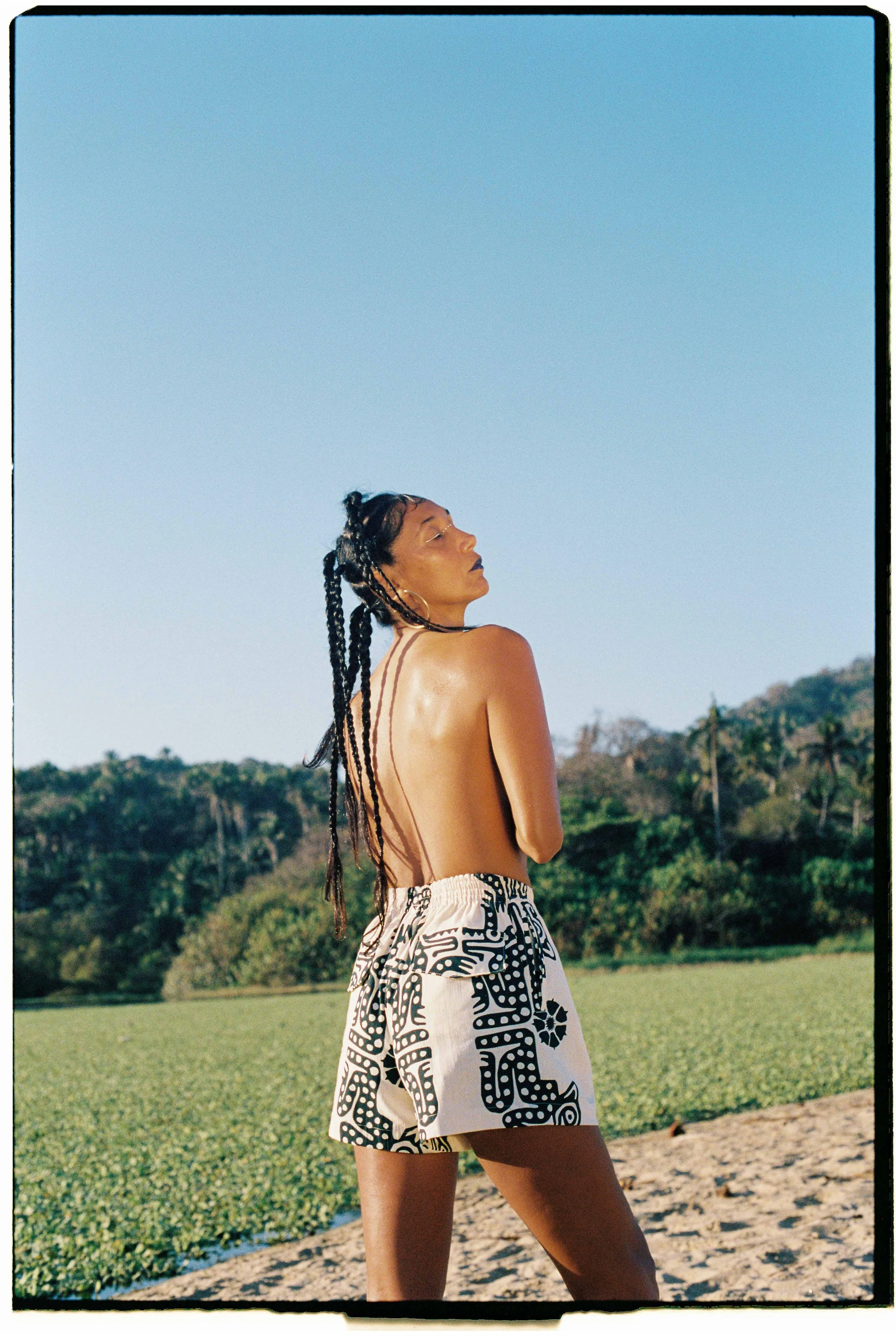 A woman with braids and hoop earrings stands topless in a natural outdoor setting with trees and a clear blue sky, wearing black and white patterned shorts.