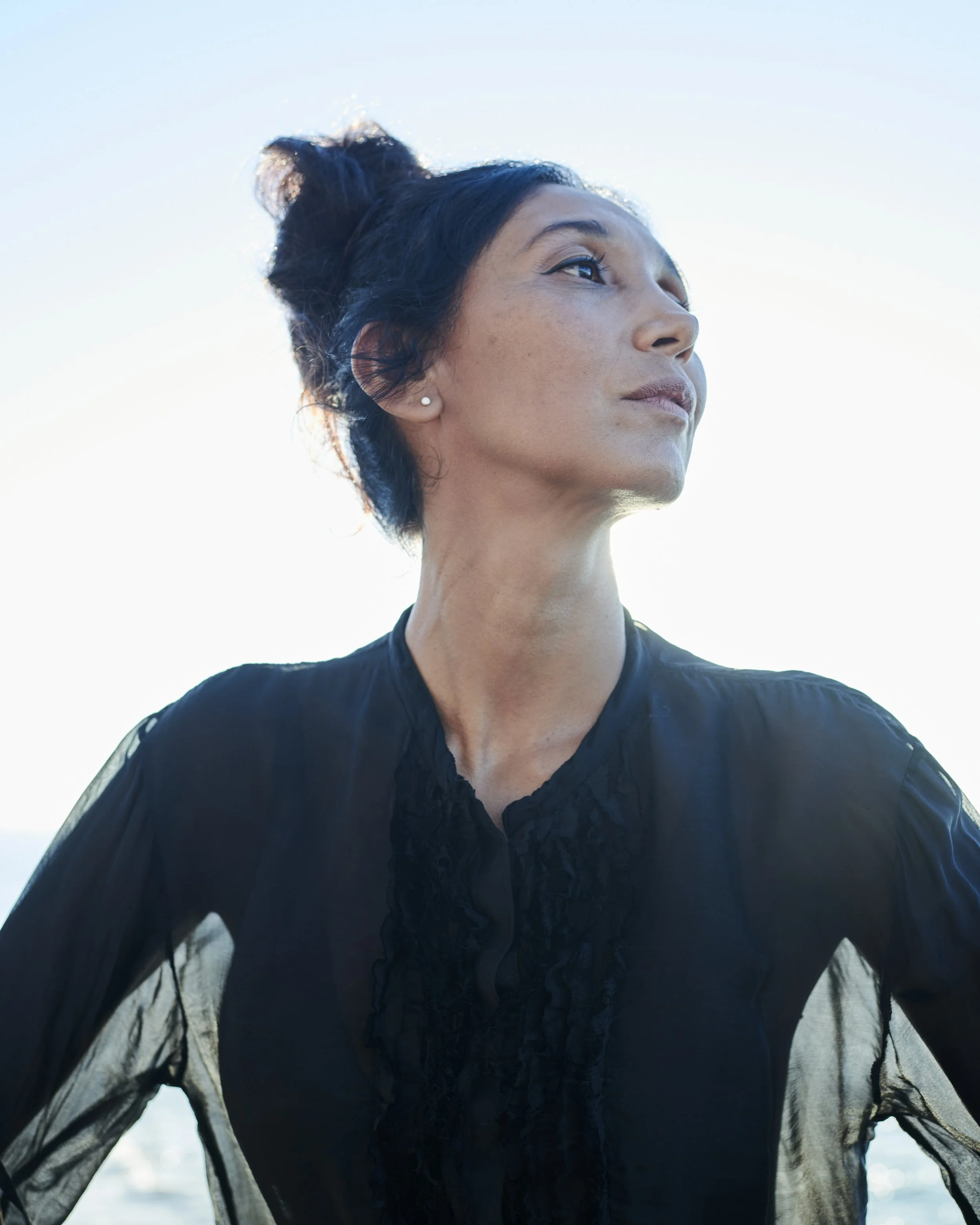 A woman with dark hair styled in a loose bun, wearing a black sheer blouse, looking confidently to the right against a bright sky background.