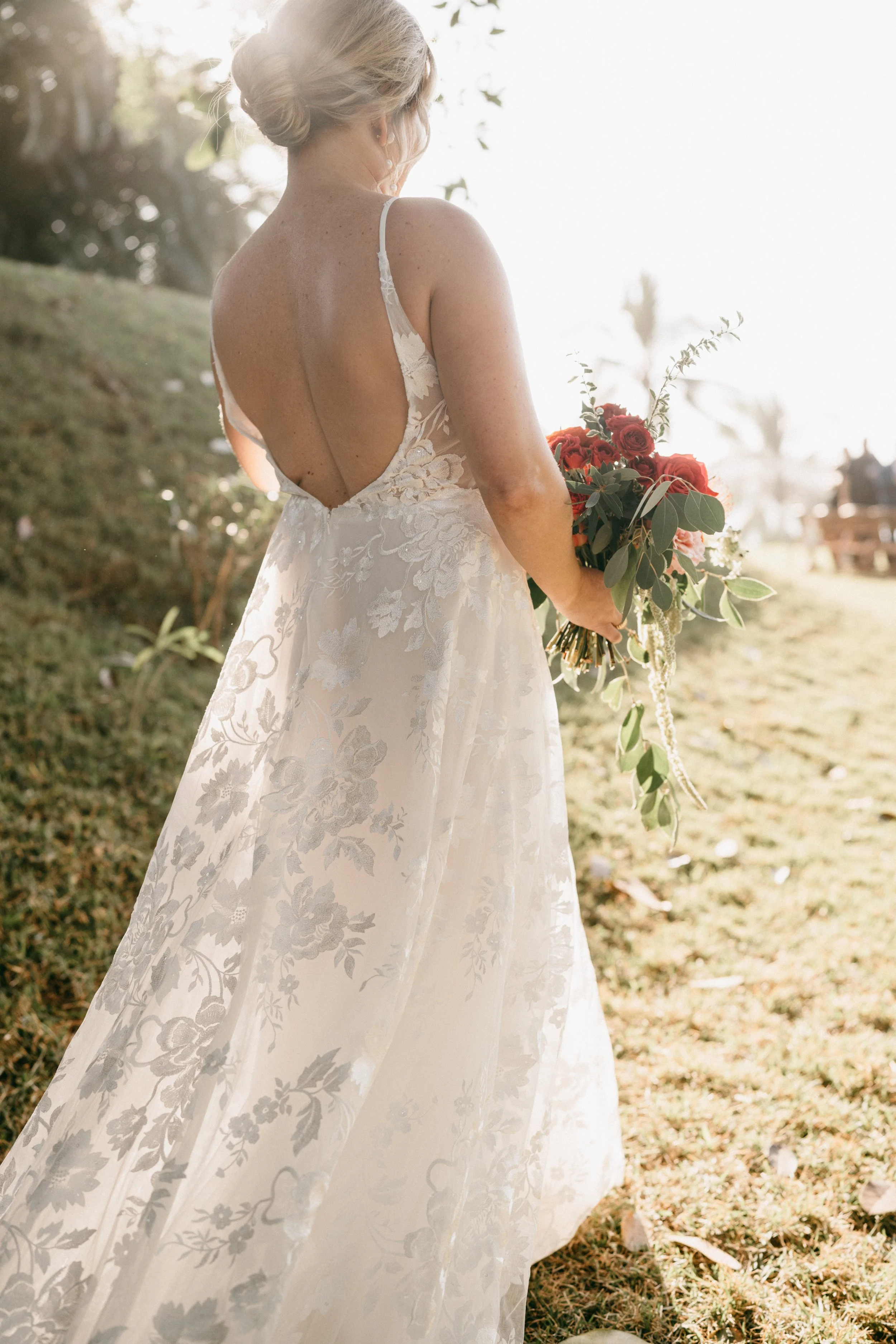 A bride in a floral lace wedding dress holding a bouquet of red and pink roses and greenery, standing outdoors on a grassy area with sunlight filtering through trees.