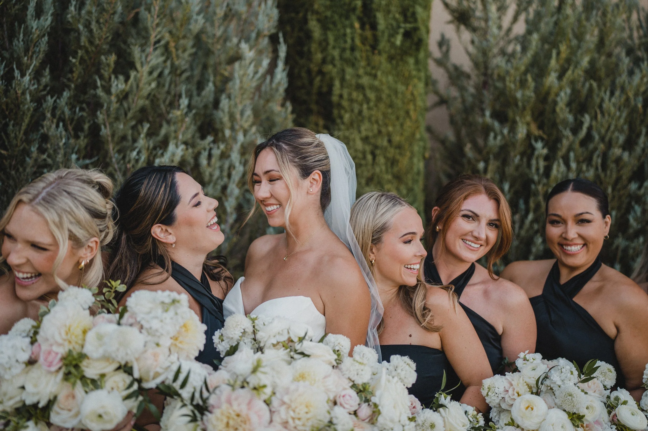 Bride and bridesmaids smiling and laughing during a wedding celebration outdoors, with lush greenery in the background.