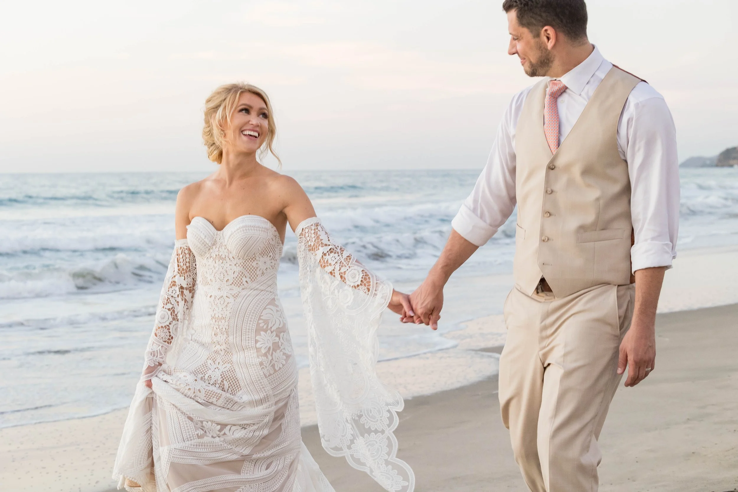 A bride and groom holding hands and walking on the beach during sunset.
