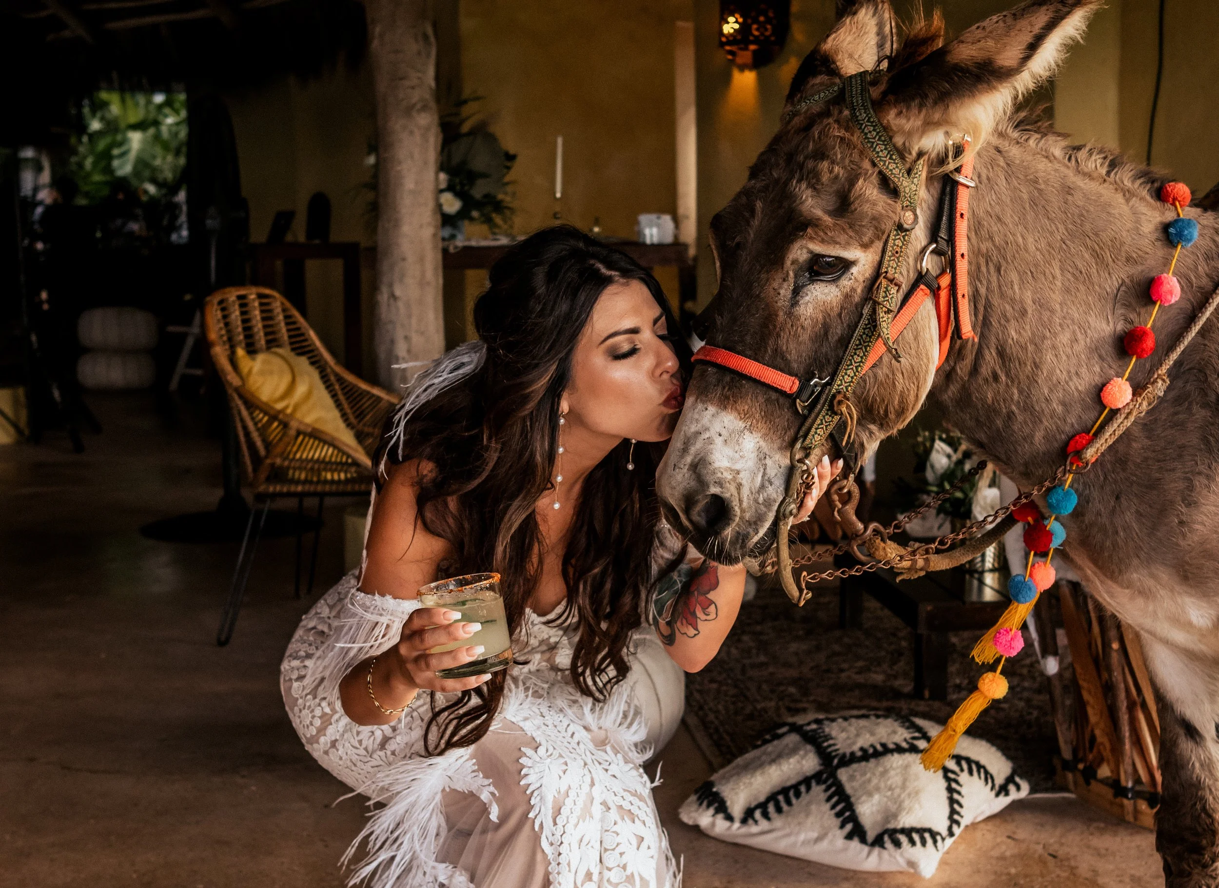 A woman with long dark hair, wearing a white lace dress, is kneeling and kissing a decorated donkey on the nose. The woman is holding a drink in her right hand, and the donkey has colorful pom-pom garland on its harness.