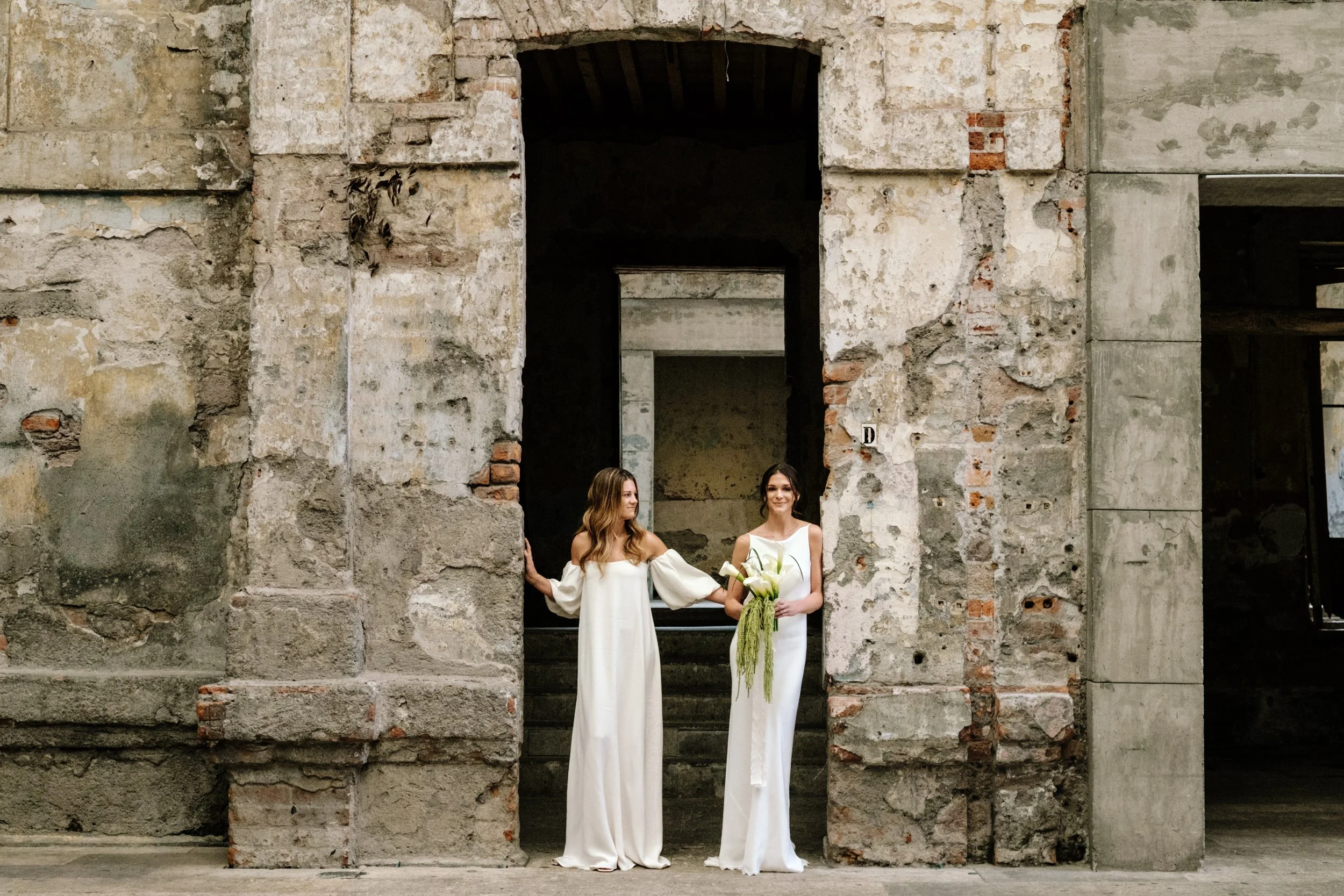 Two women in white dresses standing in front of a weathered brick and concrete wall, one holding a bouquet of white flowers.