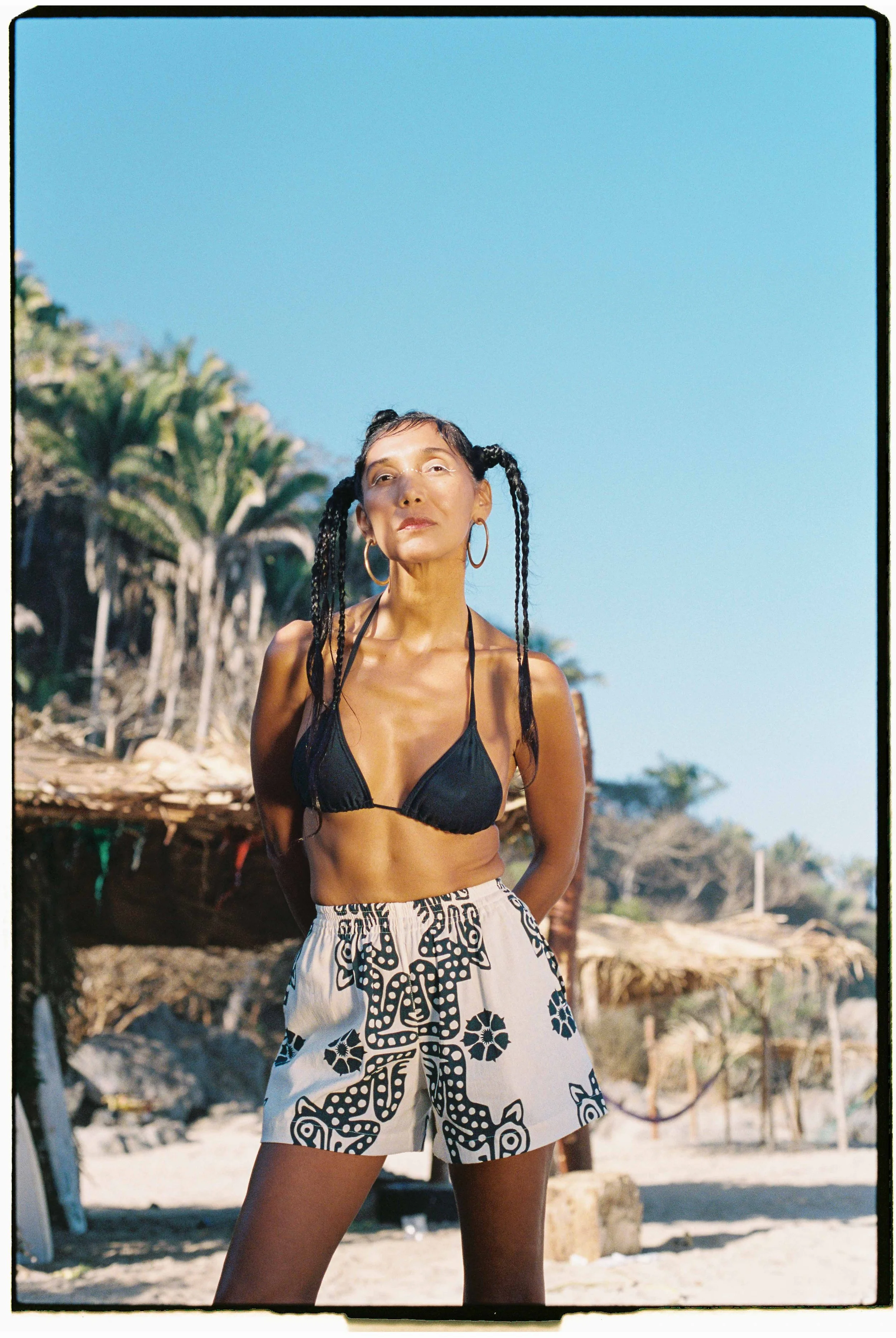 A woman in a black bikini top and patterned shorts stands outdoors on a sandy beach, with palm trees and a thatched hut in the background under a clear blue sky.