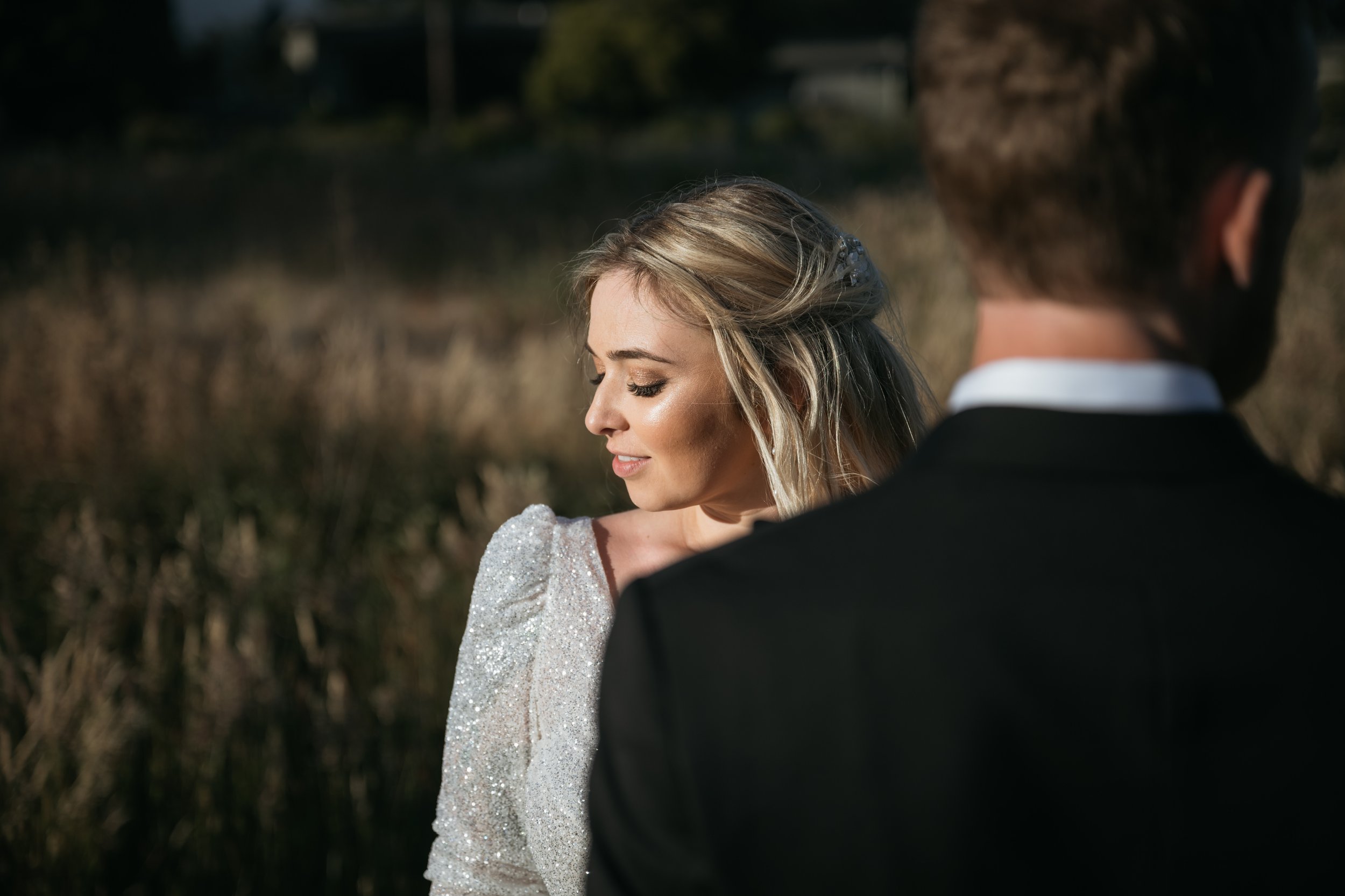 A woman in a satin dress with puffed sleeves and a man in a black suit standing outdoors in a natural setting with tall grasses and trees.