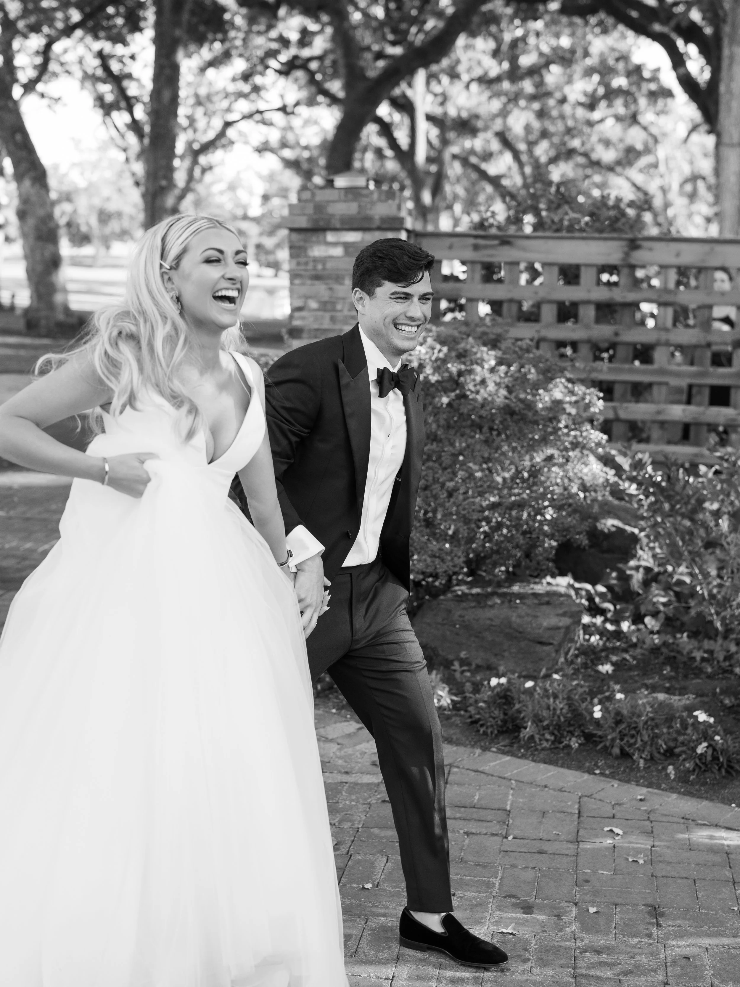 A black and white photo of a bride and groom holding hands, laughing and walking outdoors.