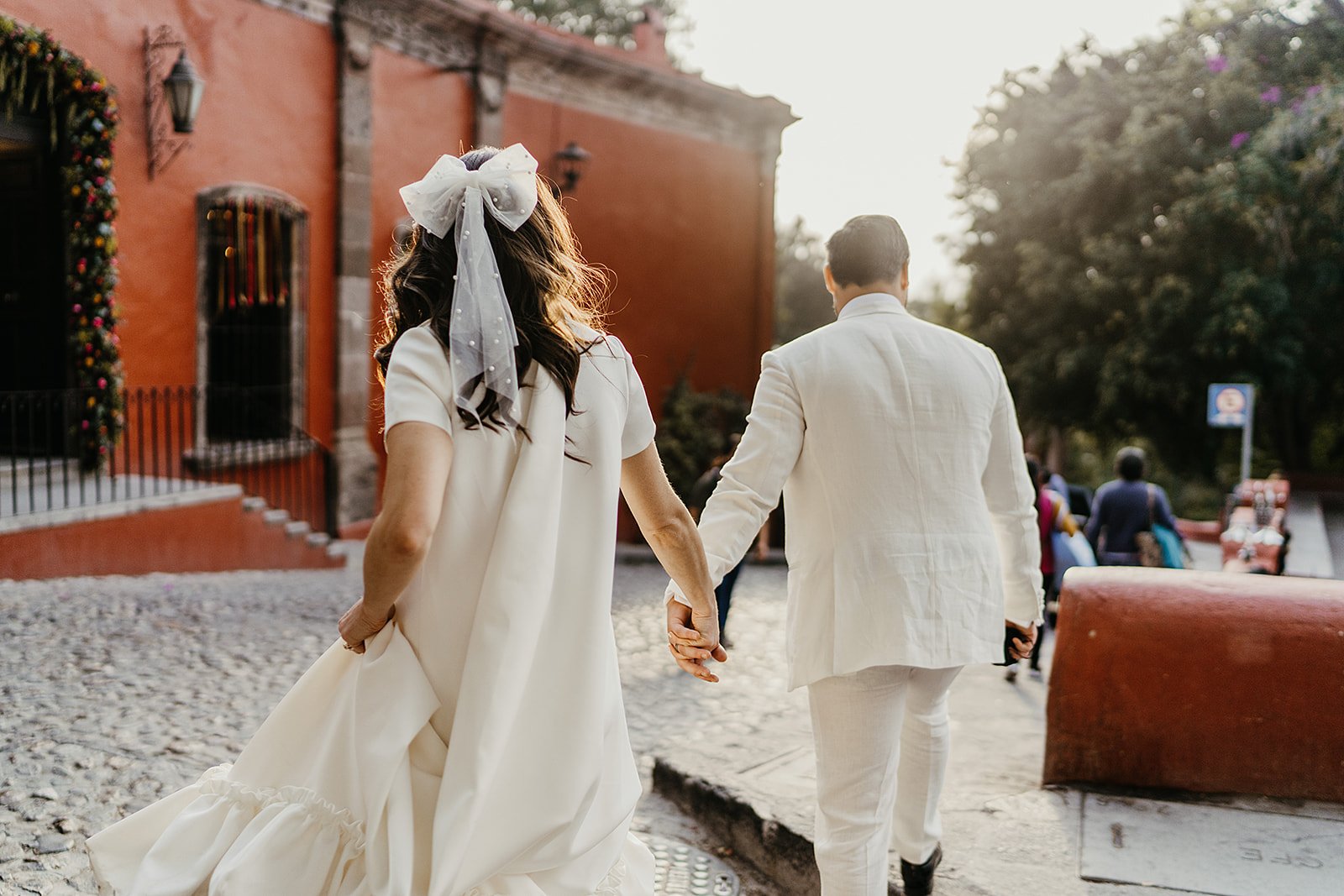 A bride and groom walking hand in hand on a cobblestone street during sunset, with friends in the background and a red building on the side.