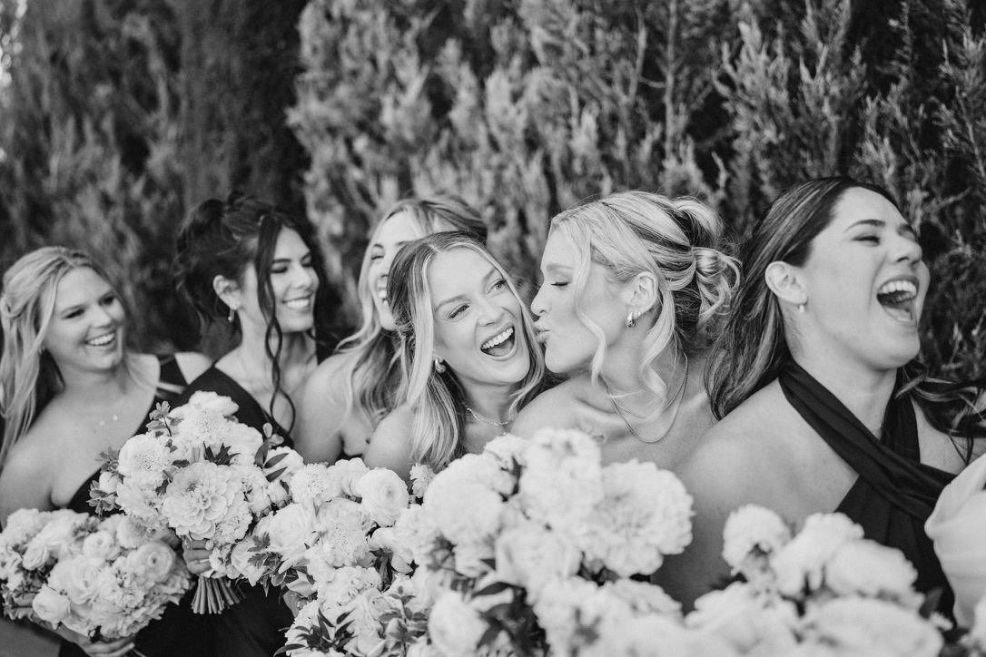 Black and white photo of a group of women in formal dresses holding bouquets, laughing and hugging outdoors.