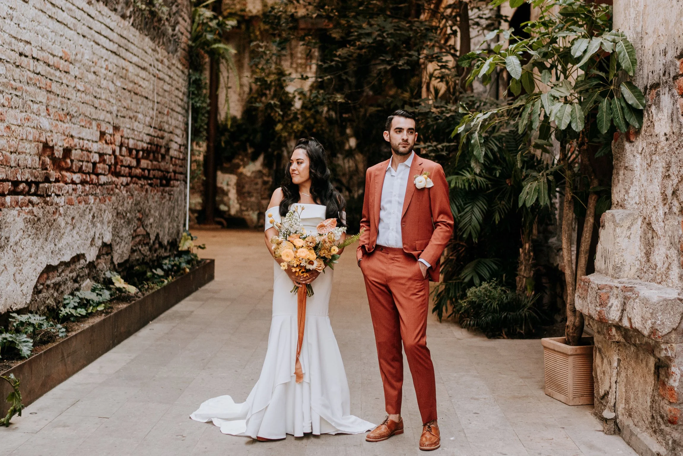 A bride in a white wedding dress holding a bouquet of flowers and a groom in a rust-colored suit standing in an outdoor garden with brick and stone walls, green plants, and trees.