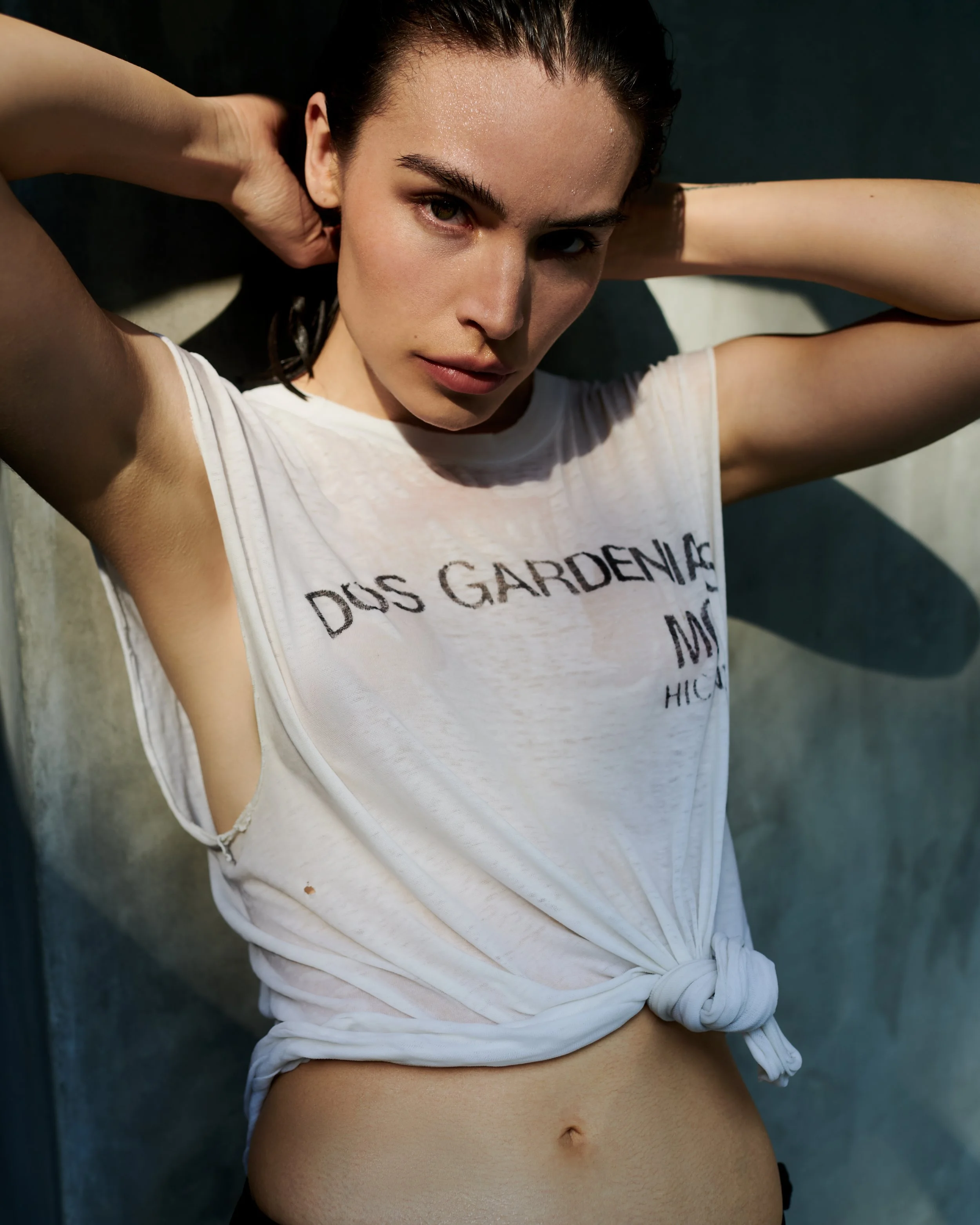A young woman with dark hair posing with her hands behind her head, wearing a white sleeveless T-shirt tied at the waist, outdoors in sunlight.