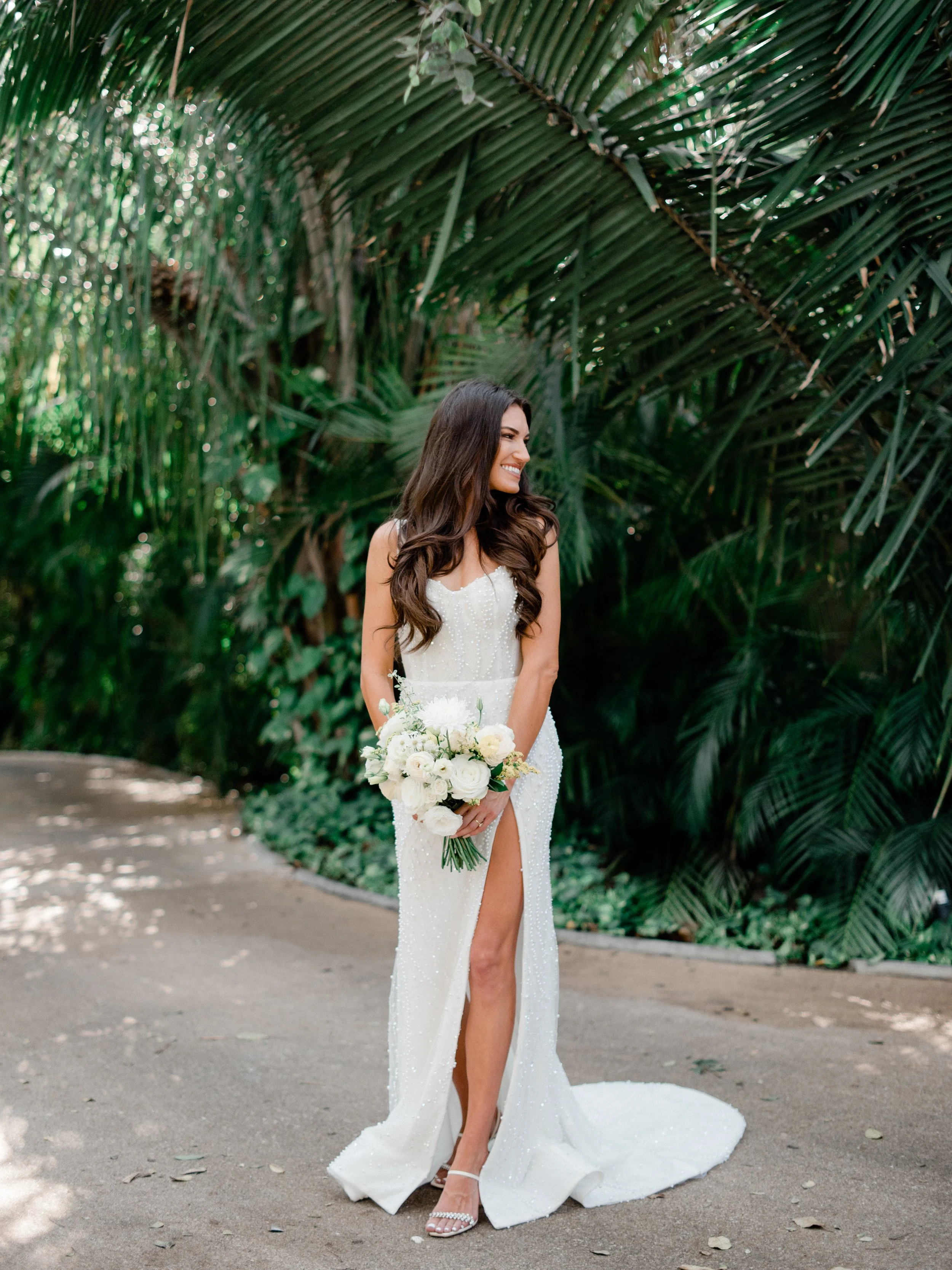 A bride in a white gown with a slit in the front, holding a bouquet of white roses, standing on a dirt path surrounded by lush green tropical foliage, smiling and looking to the side.