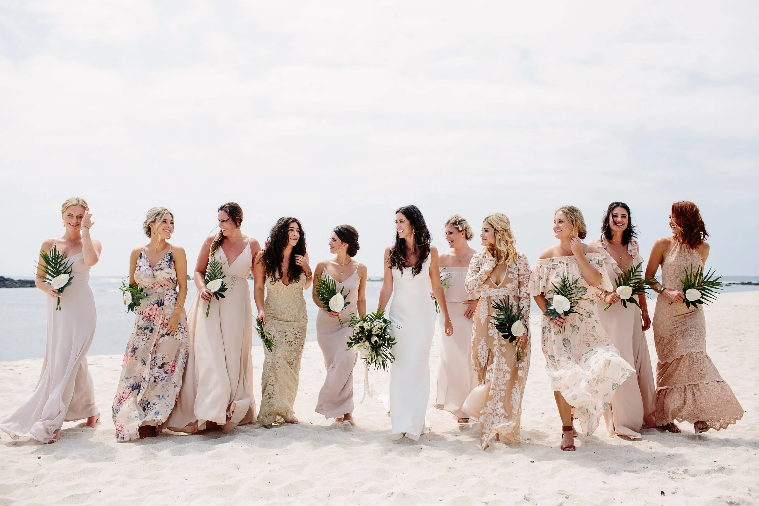 A group of women in wedding dresses walking on a beach, carrying floral bouquets, smiling and enjoying the sunny day.