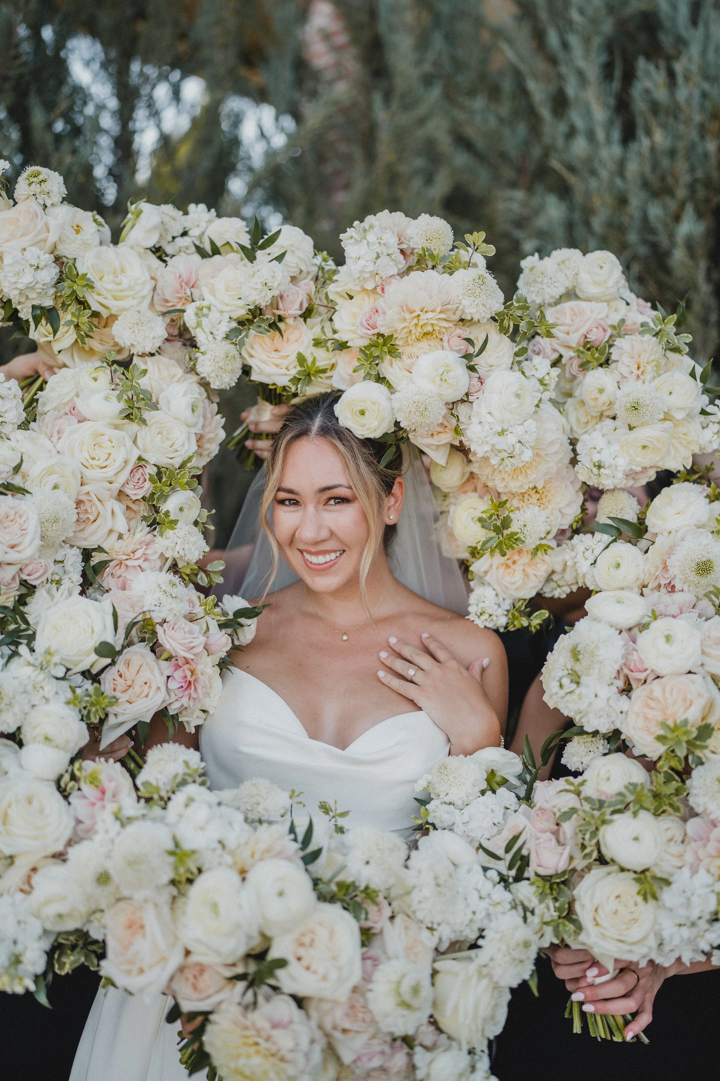 Bride smiling through a large floral arch made of white and pale pink roses and other flowers.