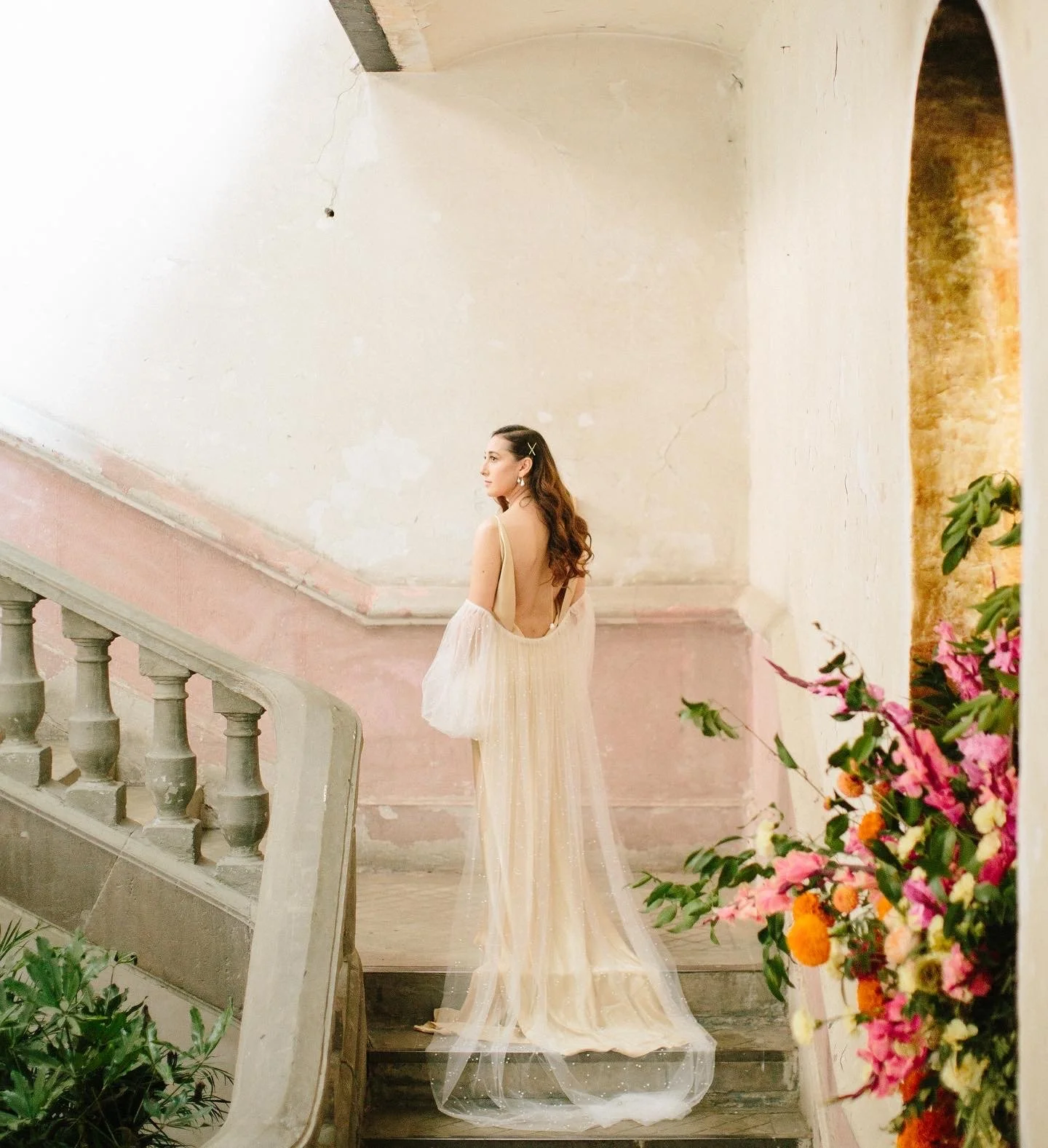 A woman in a luxurious dress standing on a staircase with colorful flowers in the foreground and a rustic wall in the background.