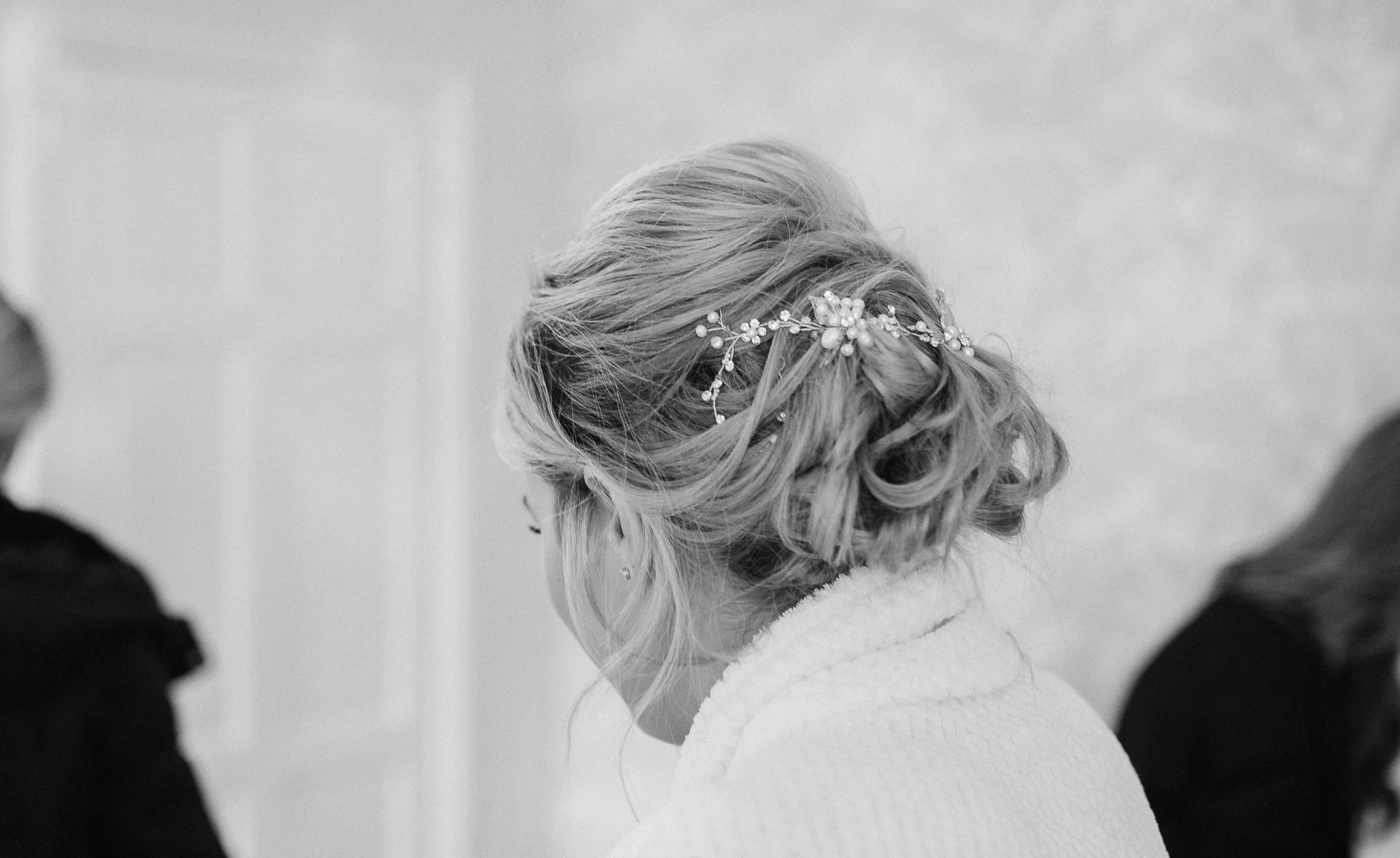 A woman with styled blonde hair wearing a decorative hairpiece, seen from behind, with two partially visible individuals on either side in the background.