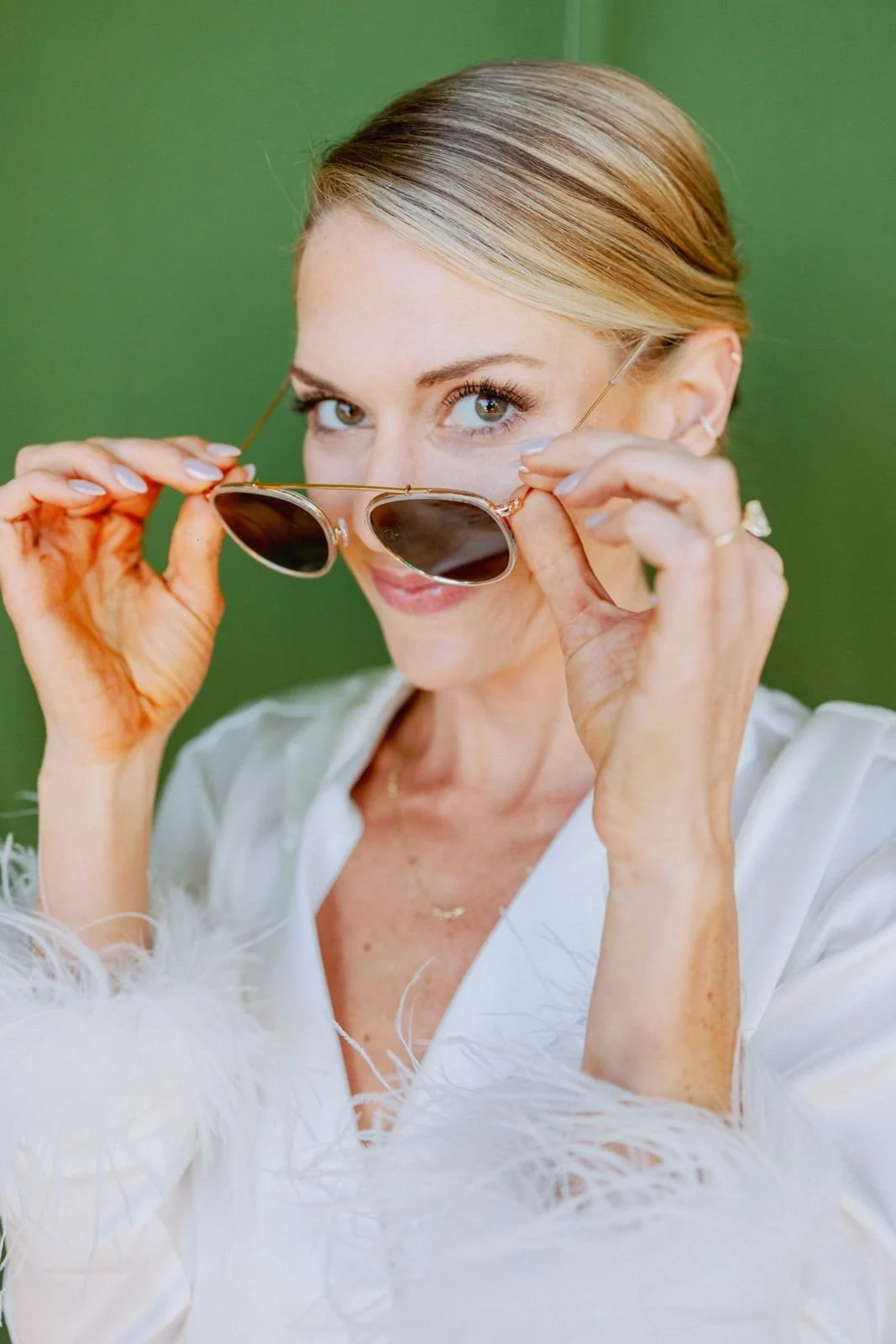 A woman with blonde hair in a sleek style, lifting sunglasses with both hands, against a green background.