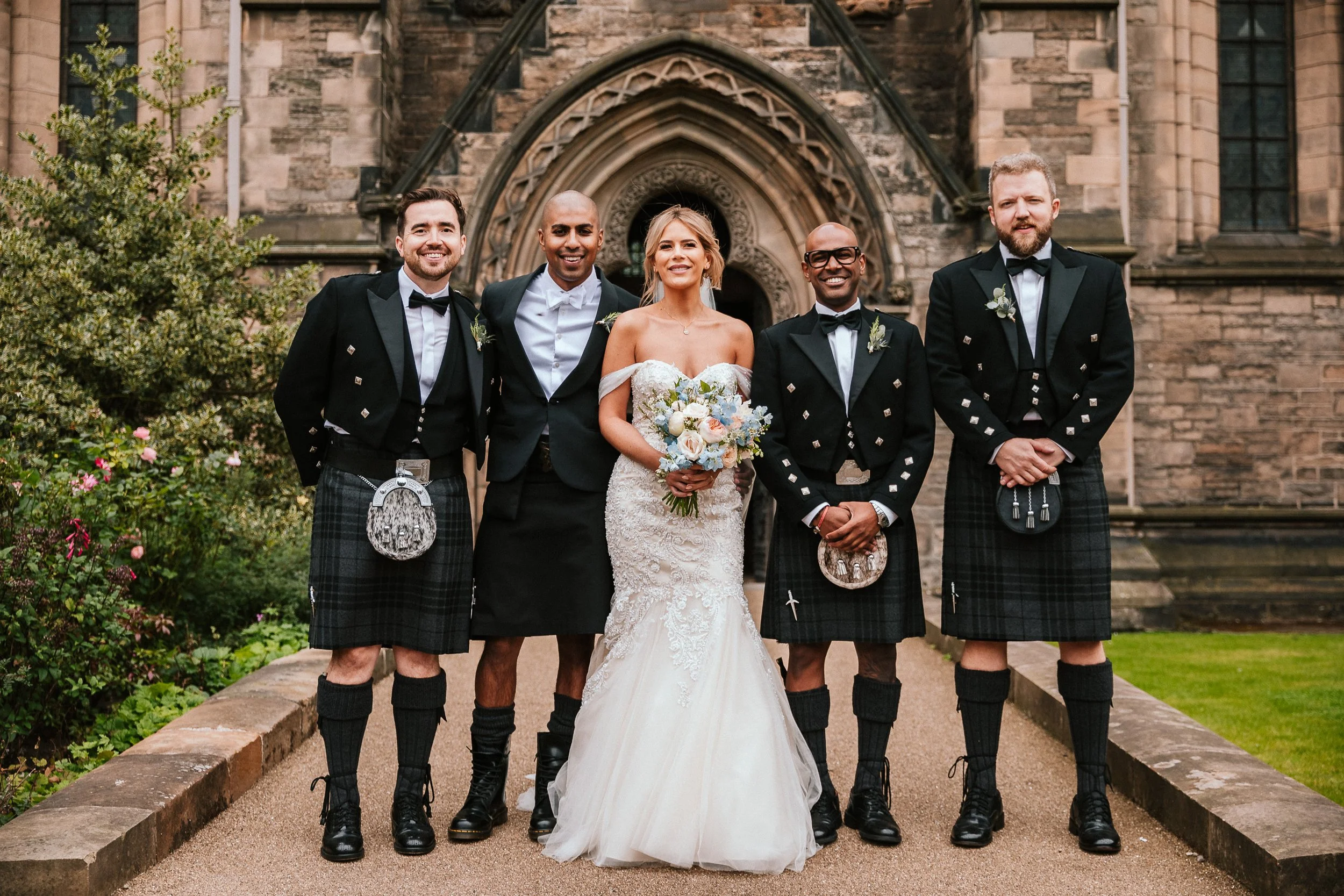 Bride in wedding dress holding bouquet, flanked by four men in traditional Scottish kilts and tuxedos, standing outside a historic stone building with gothic architectural details.