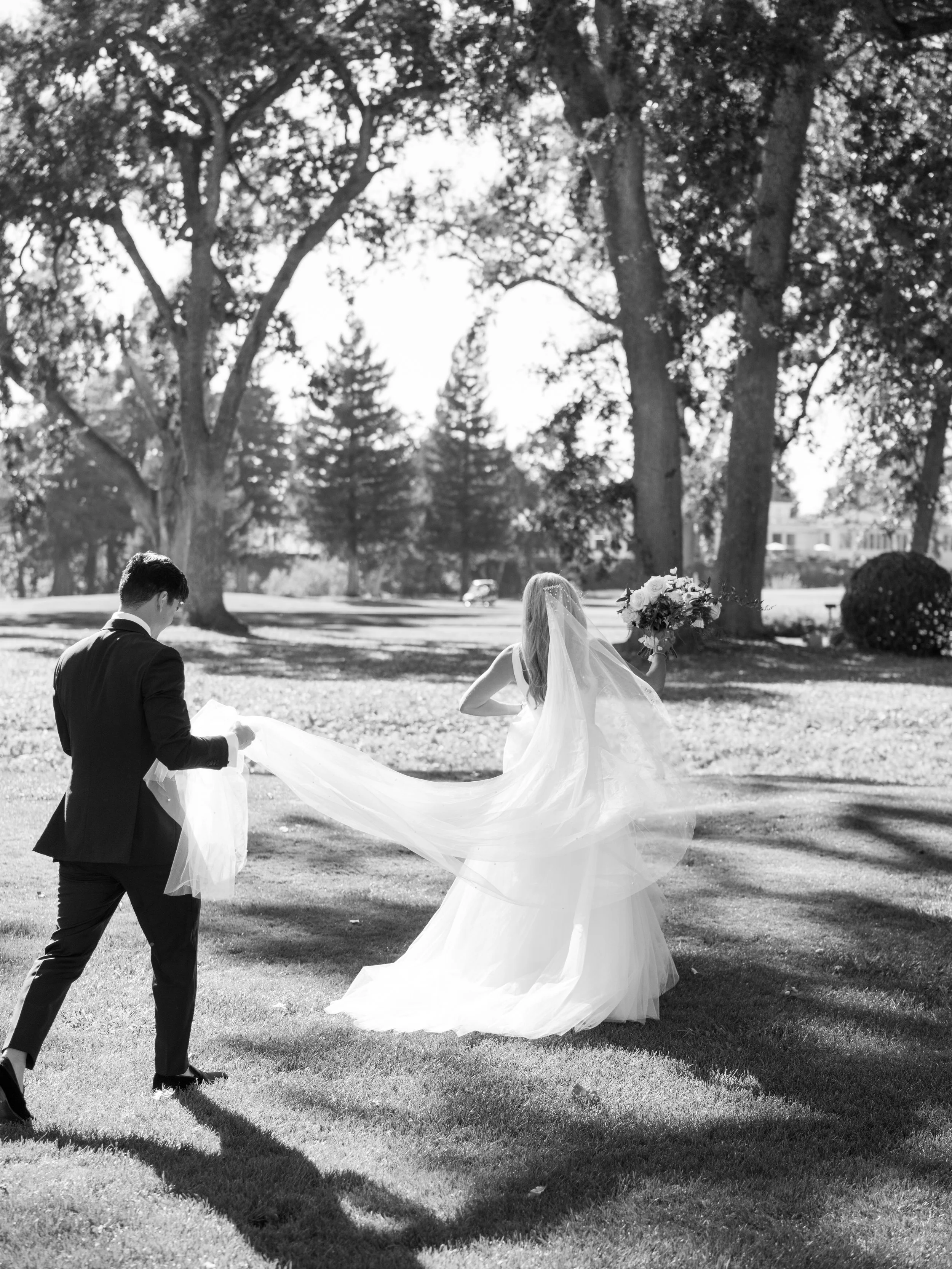 A bride and groom walking in a park, with the groom holding the bride's train and the bride holding a bouquet of flowers, surrounded by tall trees casting shadows.