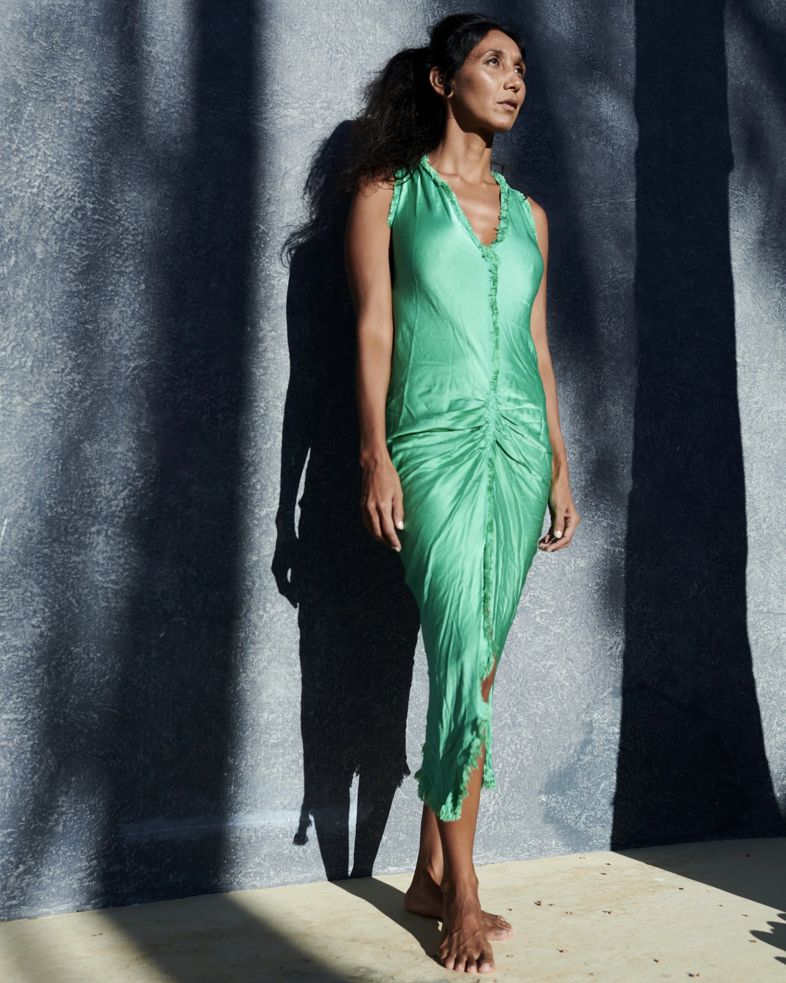 Woman in a green dress standing barefoot against a textured gray wall with shadows.