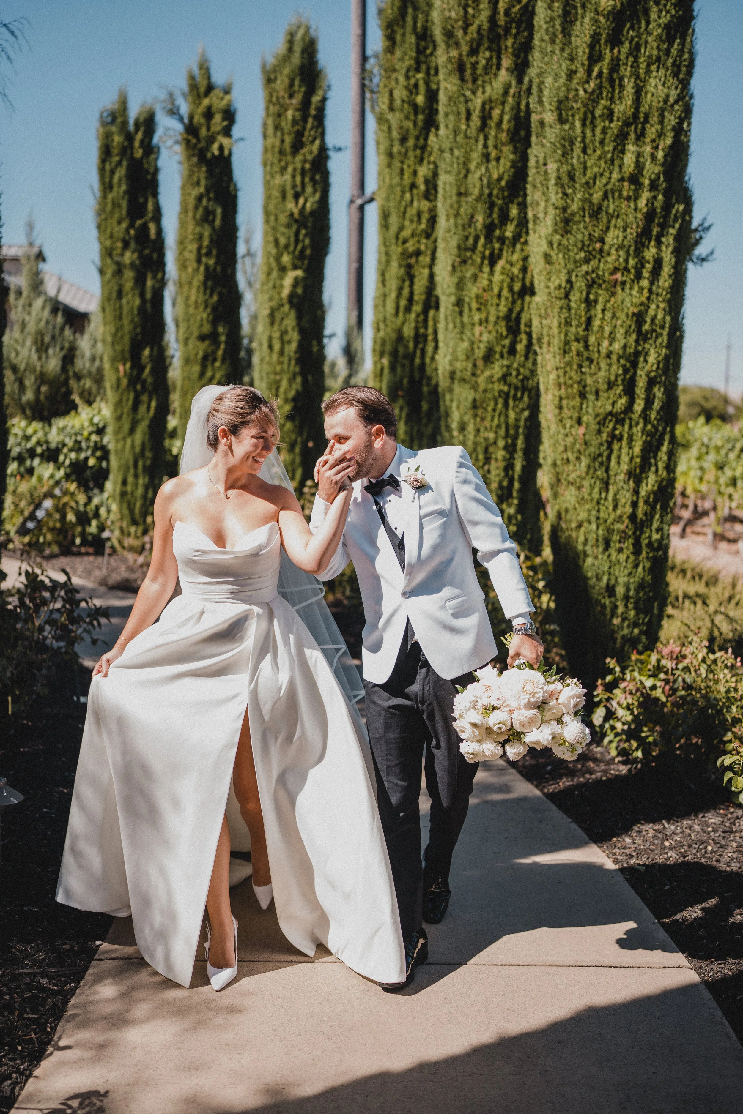 A bride and groom share a joyful moment outdoors, with the groom holding a bouquet of white flowers and the bride touching his cheek.