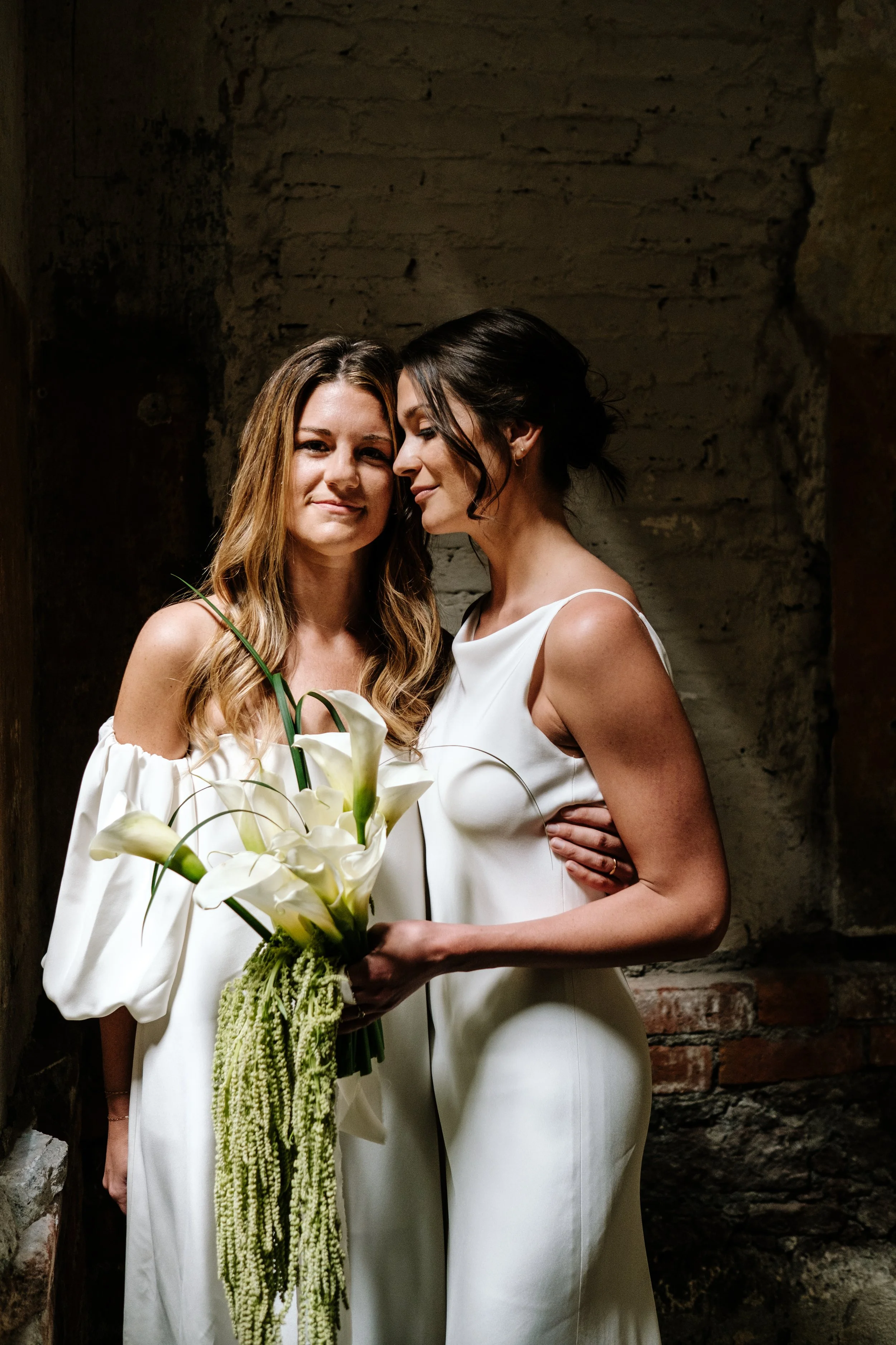 Two women in white dresses standing close together, one holding a bouquet of white calla lilies, inside a rustic brick and stone room.