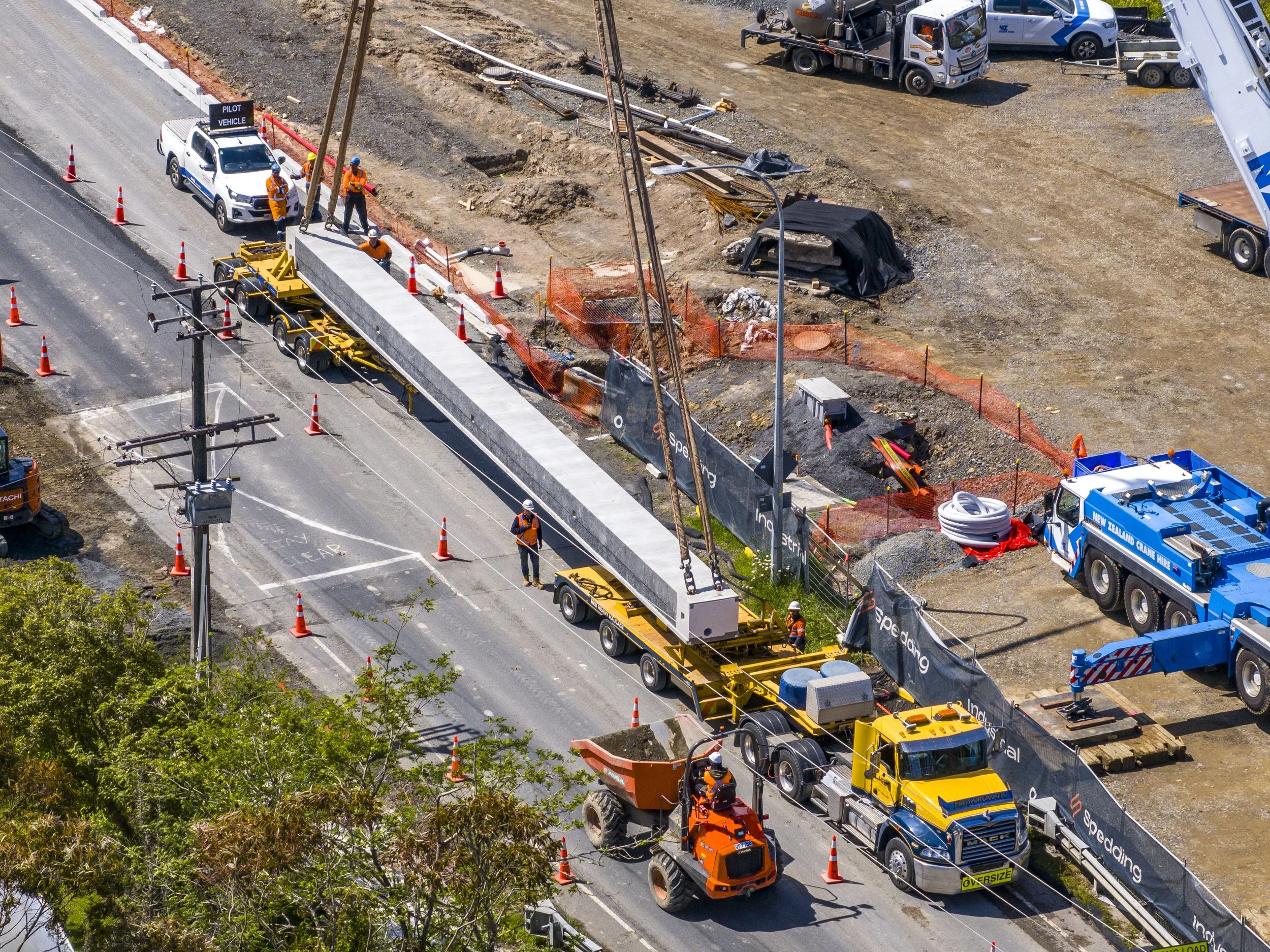 Construction workers and equipment installing a large concrete beam over a road with orange safety cones and barriers at a roadwork site.
