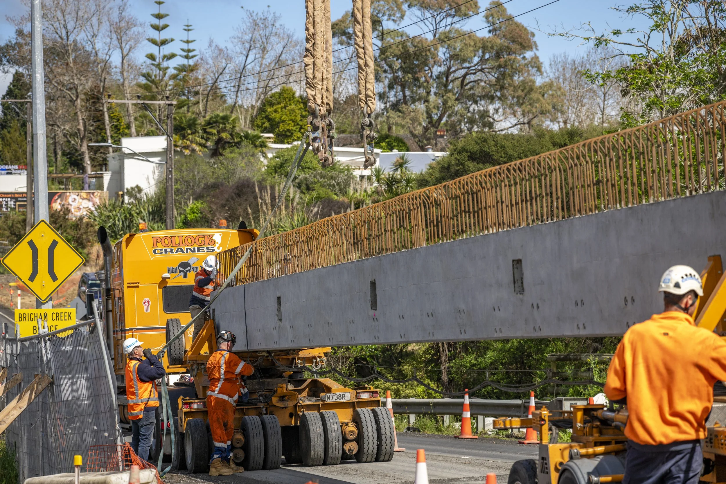 Workers with safety gear operate a large orange crane truck on a construction site near a road, lifting a long concrete barrier with steel reinforcement on top, with orange cones marking the area and a yellow road sign indicating a narrow road.
