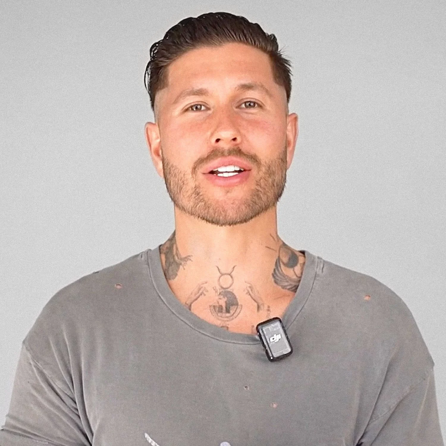 A man with short dark hair and a beard, wearing a gray shirt with small holes, standing against a plain light gray background. The man has visible tattoos on his neck and chest and is speaking.