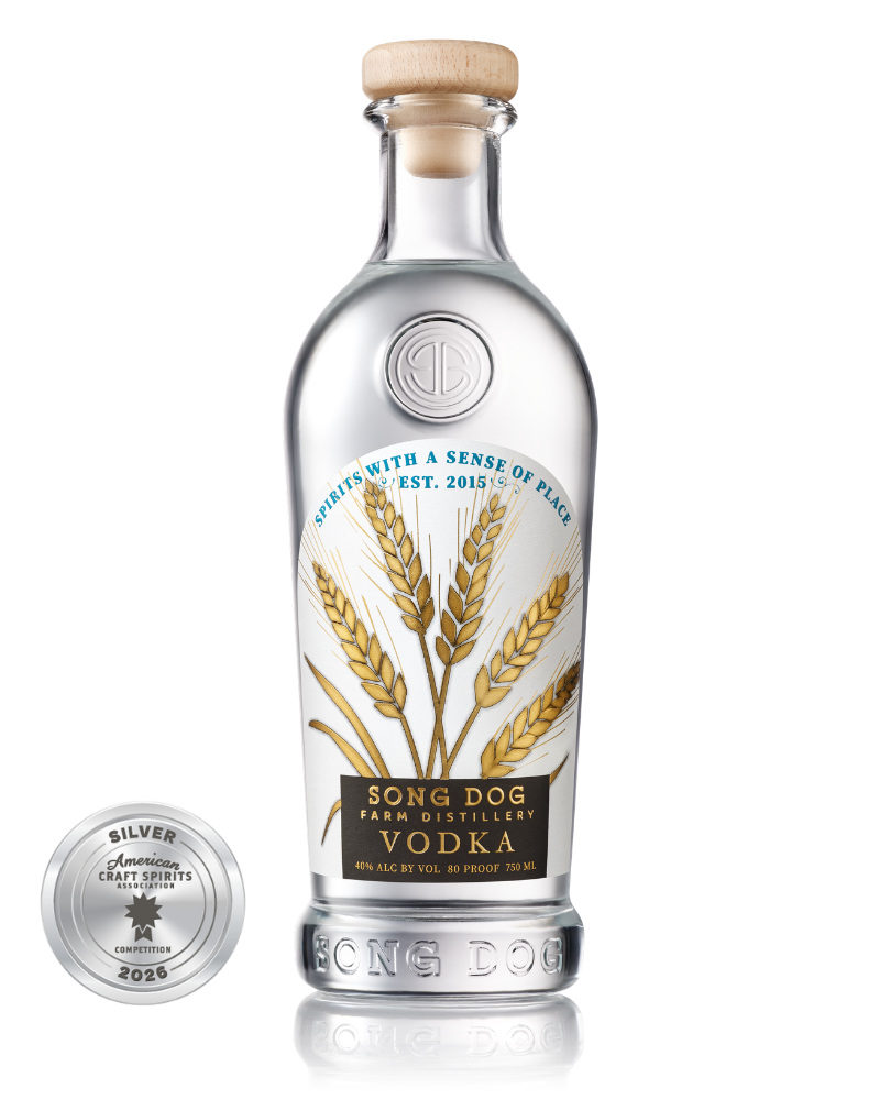 A clear vodka bottle labeled 'Song Dog Farm Distillery Vodka' with illustrations of wheat and a black label, along with a silver award medallion from the American Craft Spirits Association competition in 2026.