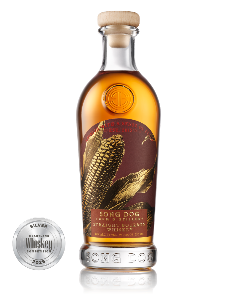 A glass bottle of Song Dog Straight Bourbon Whiskey with a wooden cork top, featuring an label with an ear of corn and leaf illustrations, and a silver Heartland Whiskey Competition 2025 silver medal to the left of the bottle.