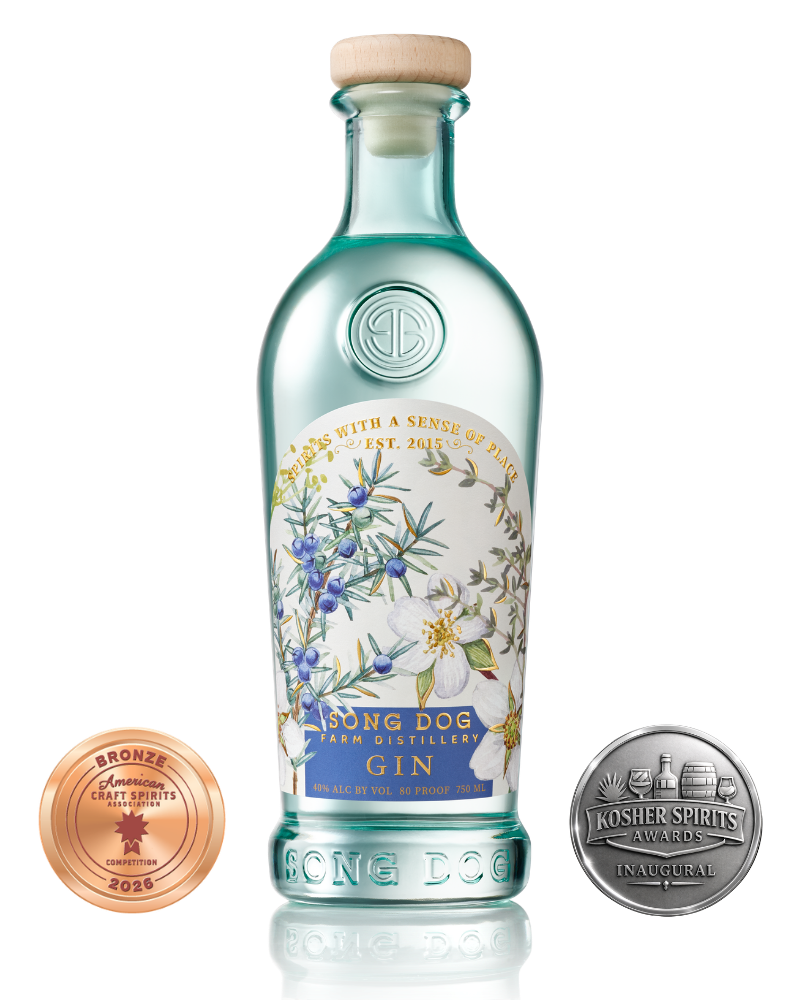 A bottle of Song Dog Farm Distillery gin, with a floral label and a cork stopper. Two award medals are positioned beside the bottle, one bronze from the American Craft Spirits Association 2026 competition, and one silver from the Kosher Spirits Awards inaugural event.