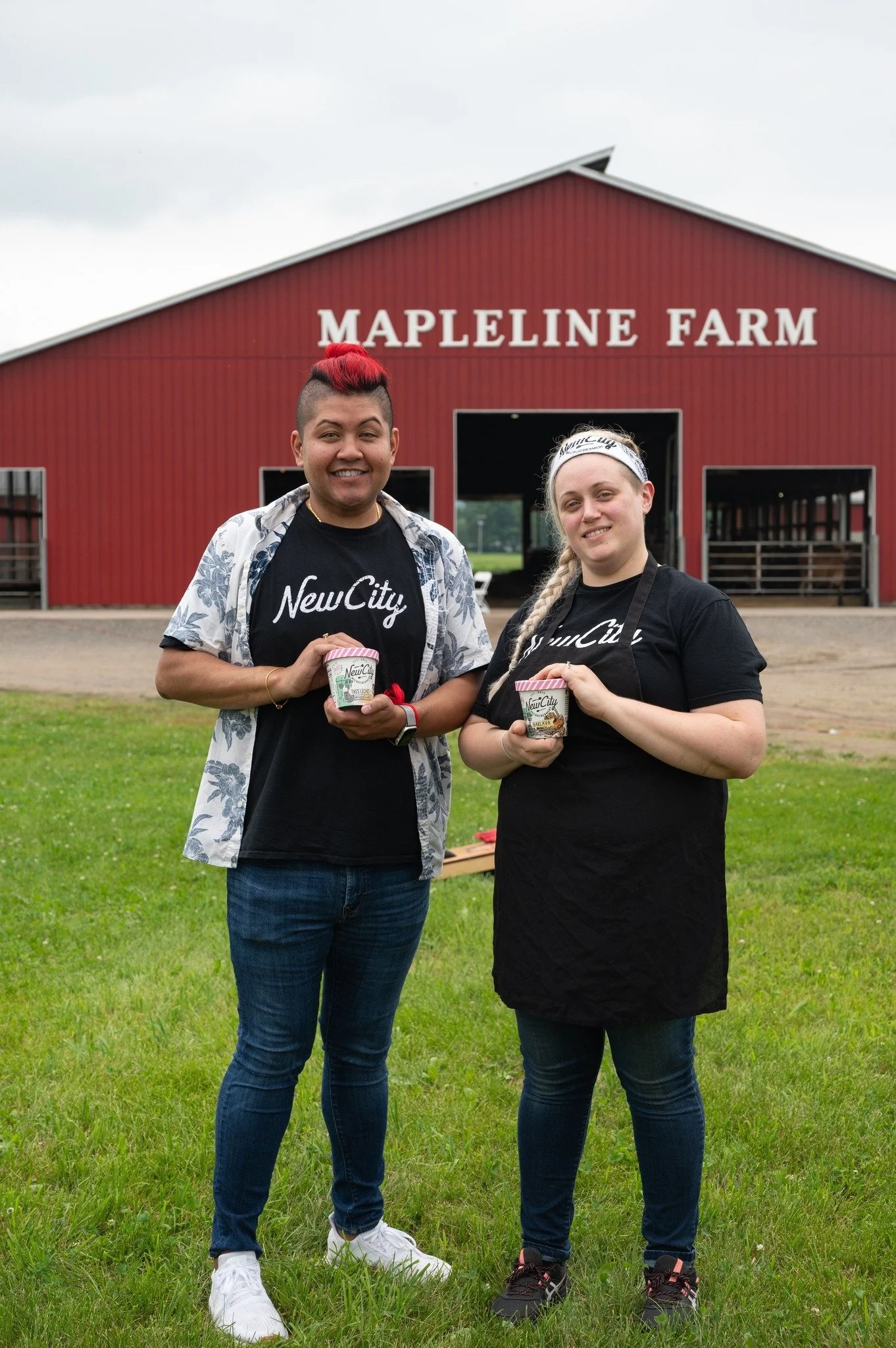 Two people standing in front of a barn with the sign Mapleline Farm on the front, holding pints of ice cream