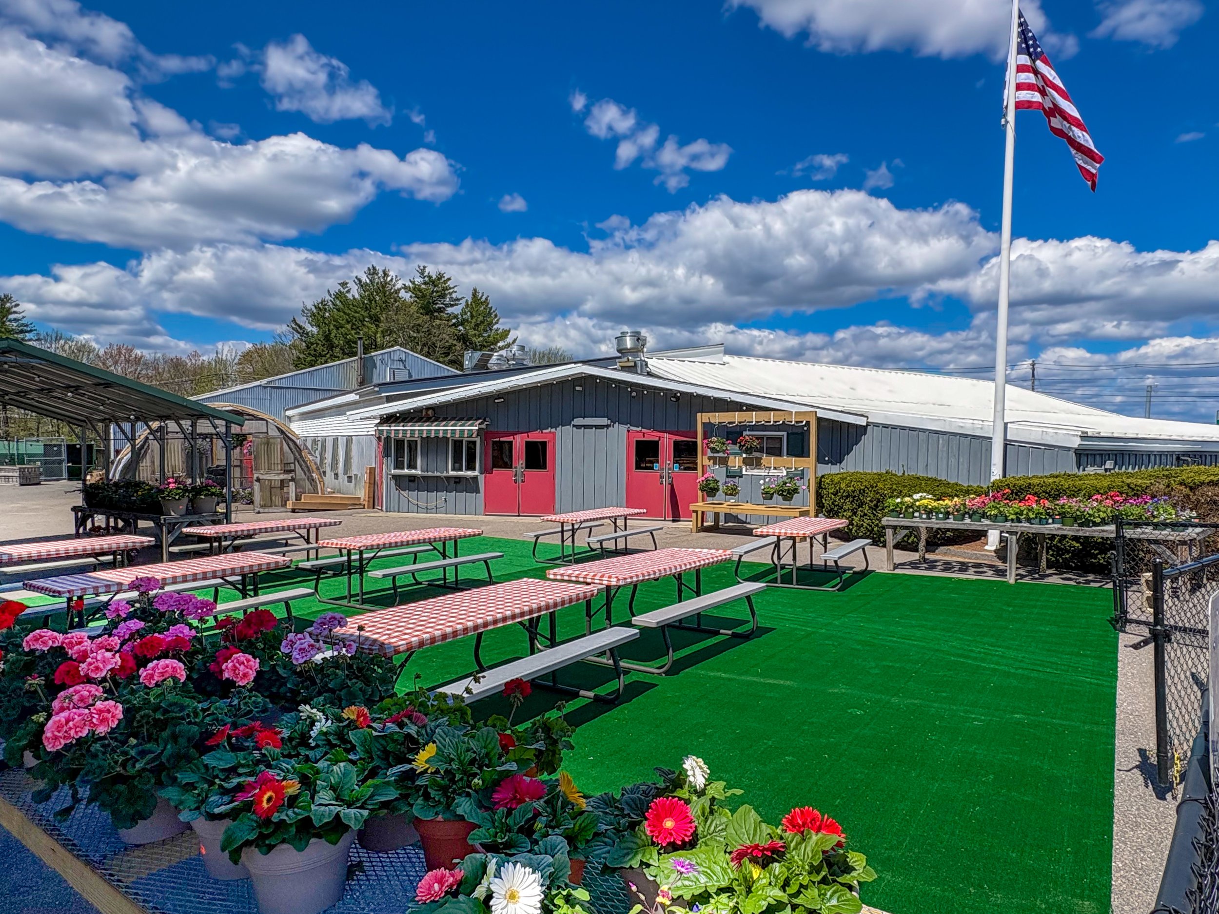 Outdoor scene with a garden center featuring colorful flowers in pots, picnic tables with red and white checkered tablecloths, a small building with red doors, and an American flag flying on a tall pole under a partly cloudy sky.