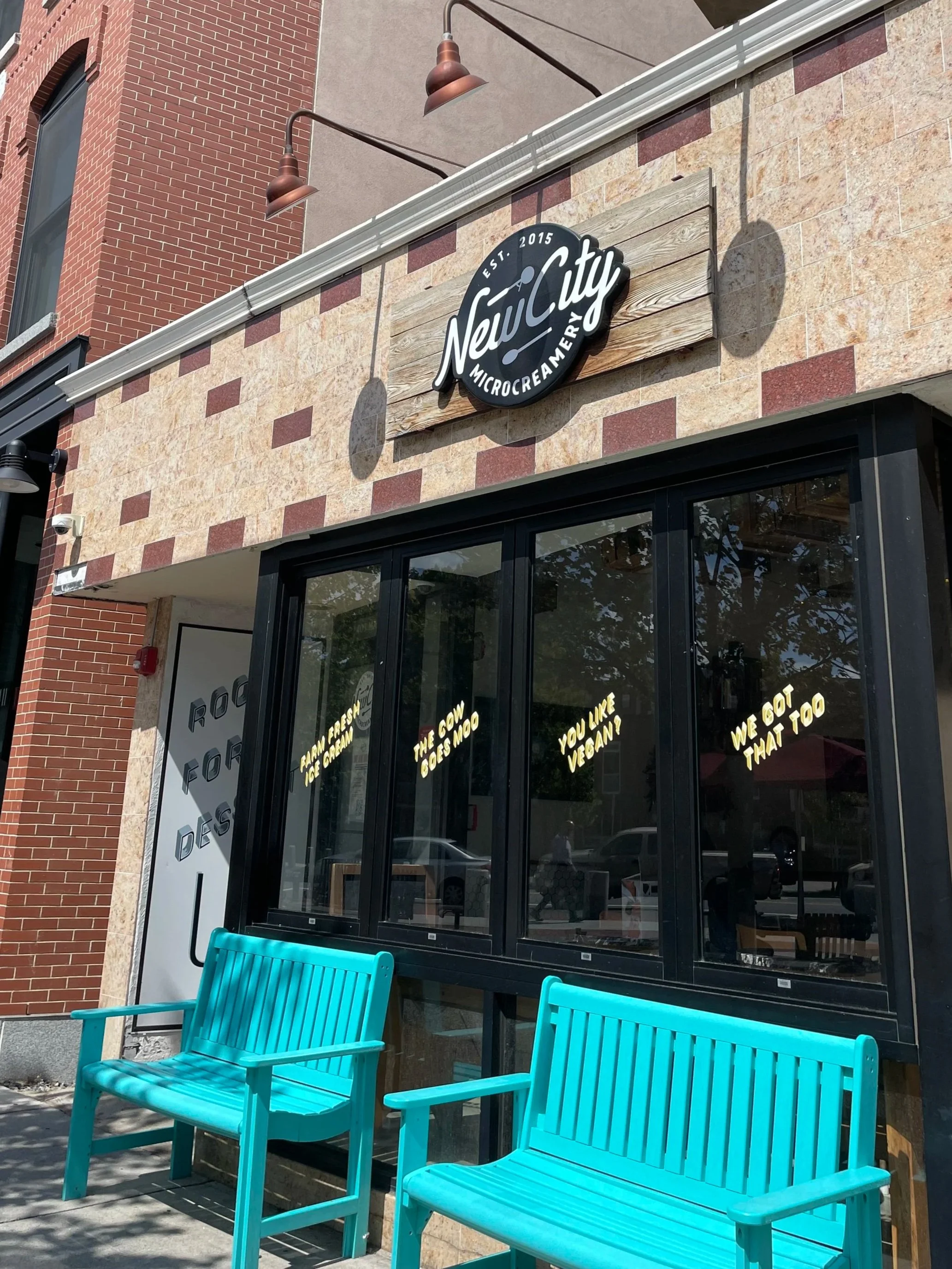 Exterior of New City Microcreamery with two bright blue benches outside, a sign on the window reading 'You like vegan? We got that too,' and a larger sign on the building face with the microcreamery's name.