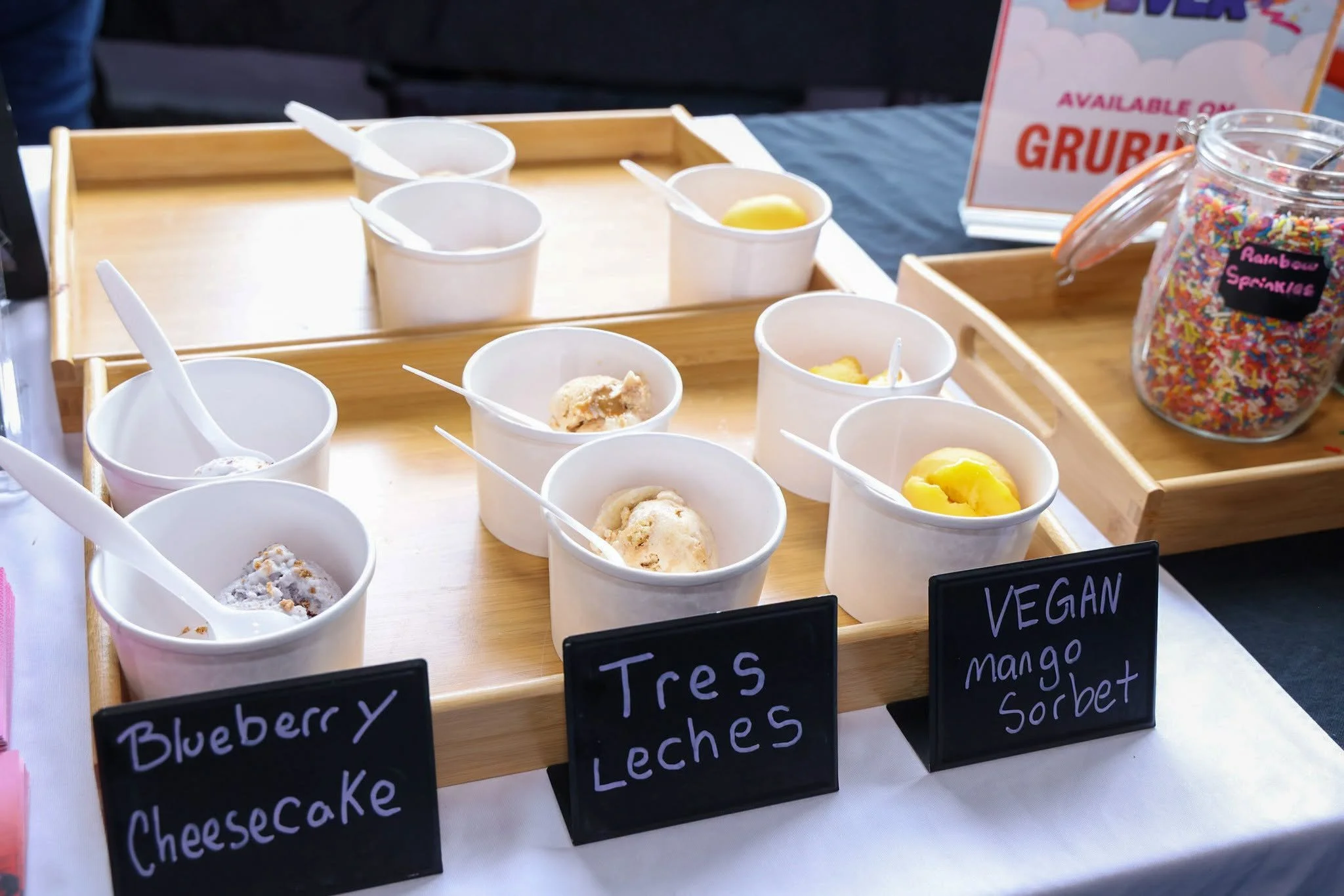 A display of multiple small cups of vegan sorbet flavors on a wooden tray, labeled with chalkboard signs including blueberry, cheesecake, and mango. A jar of rainbow sprinkles is nearby, and a sign indicates the flavors are available.