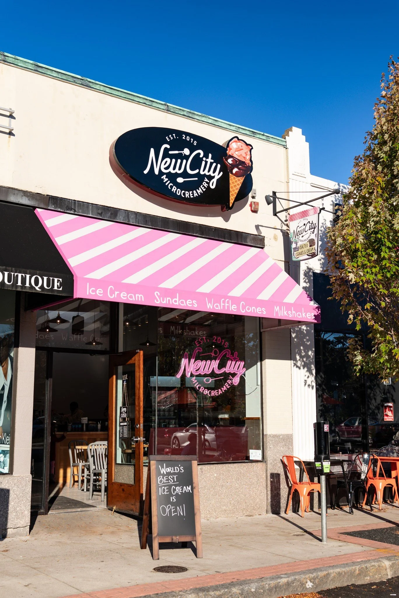Exterior of New City Micro Creamery with pink and white striped awning advertising ice cream, sundaes, waffle cones, and milkshakes, featuring a sign with pink neon text and ice cream cone graphic, with outdoor seating and sidewalk.
