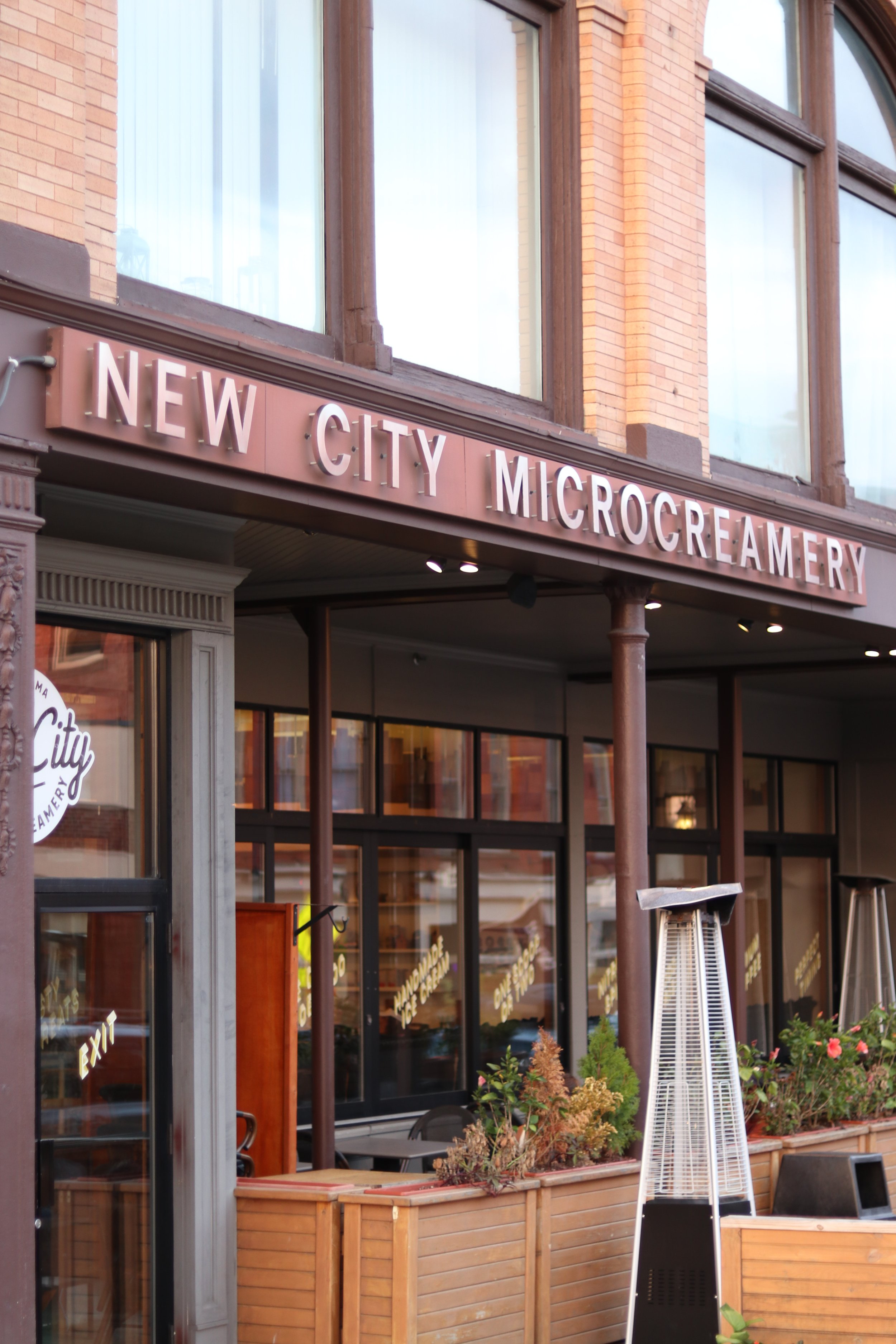 Front of a building with a sign that reads 'New City Microcreamery', featuring large windows and outdoor seating with plants and a tall outdoor heater.
