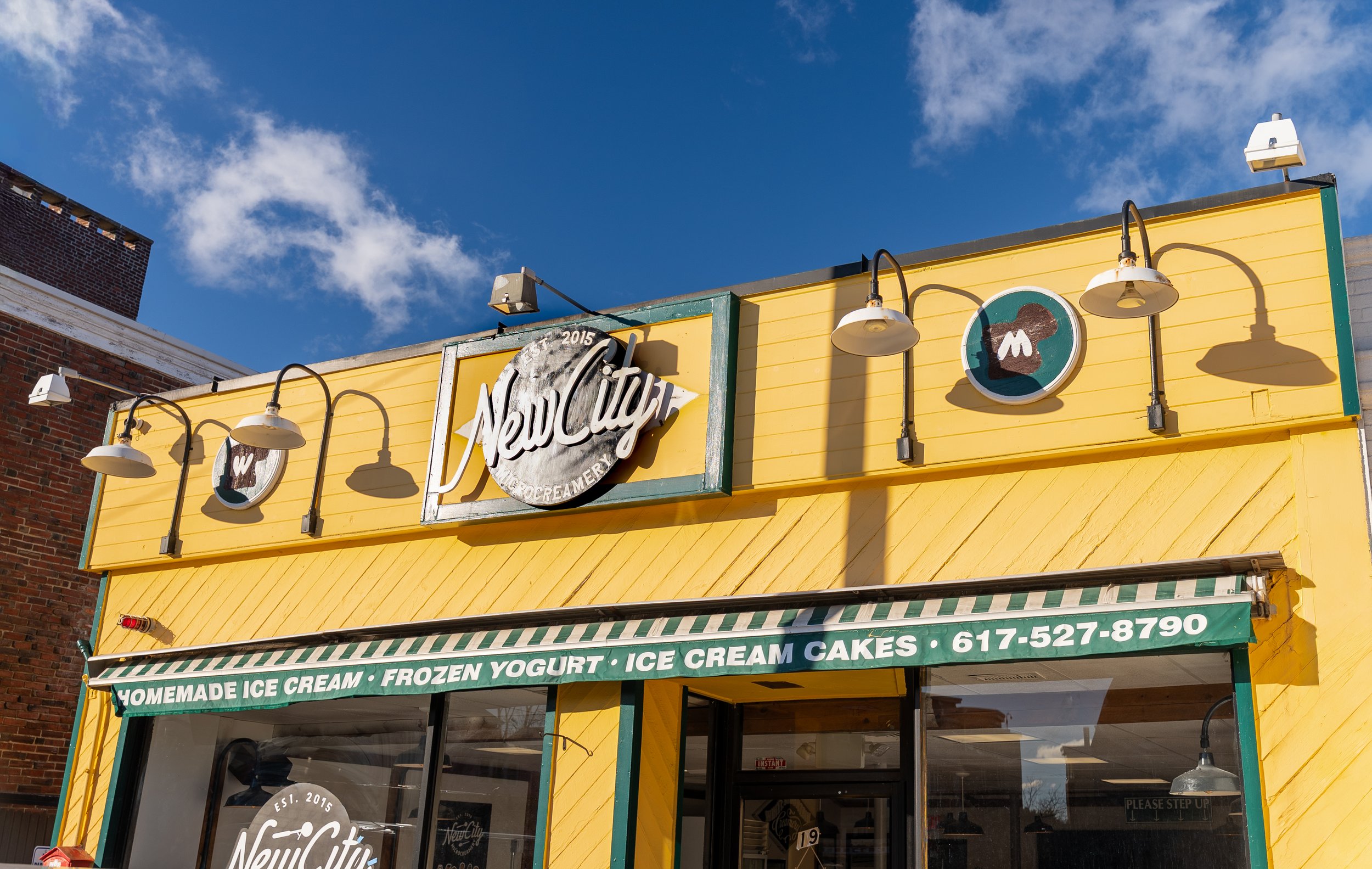 Yellow storefront of New City ice cream shop with green and white striped awning, signs advertising homemade ice cream, frozen yogurt, and ice cream cakes, and a phone number, under a partly cloudy blue sky.