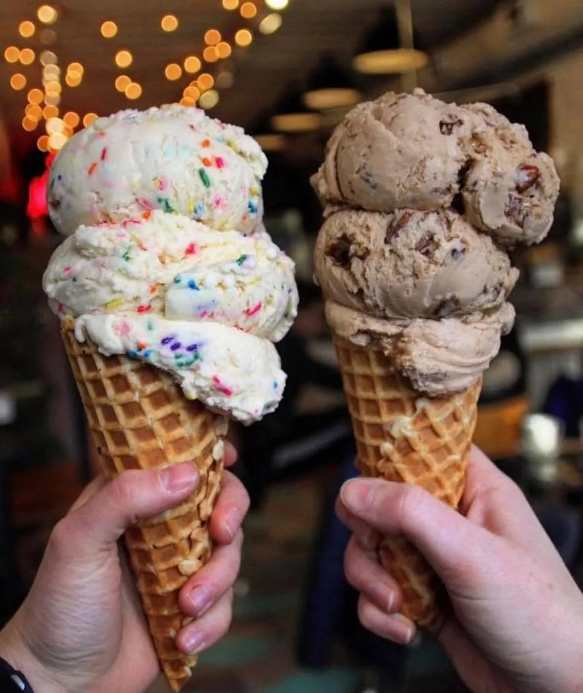 Two hands holding waffle cones with scoops of ice cream, one with colorful sprinkles and the other with chocolate chunks, against a blurred background with warm lighting.