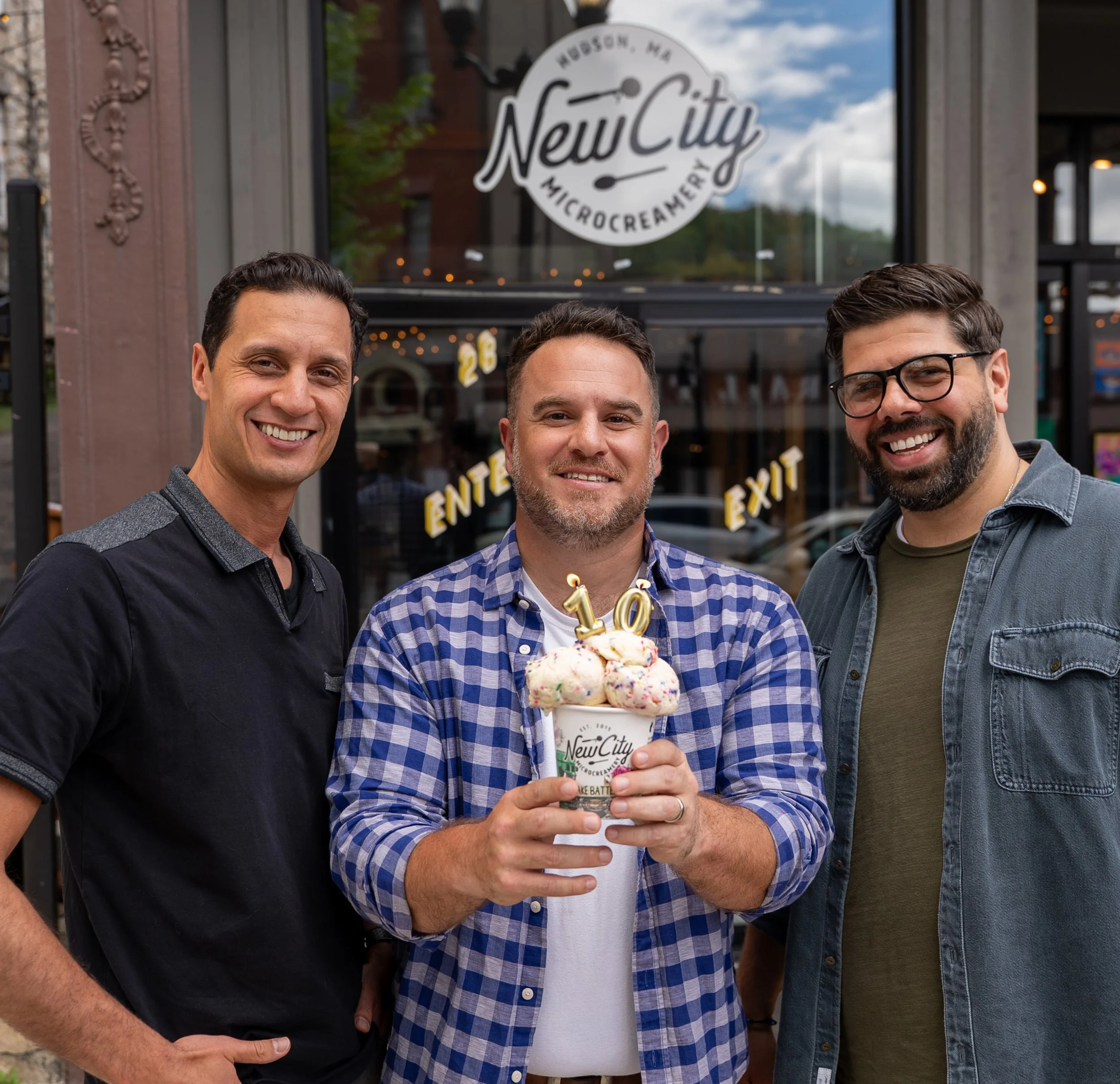 Three smiling men standing outdoors in front of a bakery or cafe with a sign that reads "New City Microcreamery." The man in the middle is holding a large ice cream sundae with a birthday candle shaped like the number 10. The men are dressed casually, and the background shows a building with large windows, some trees, and a cloudy sky.