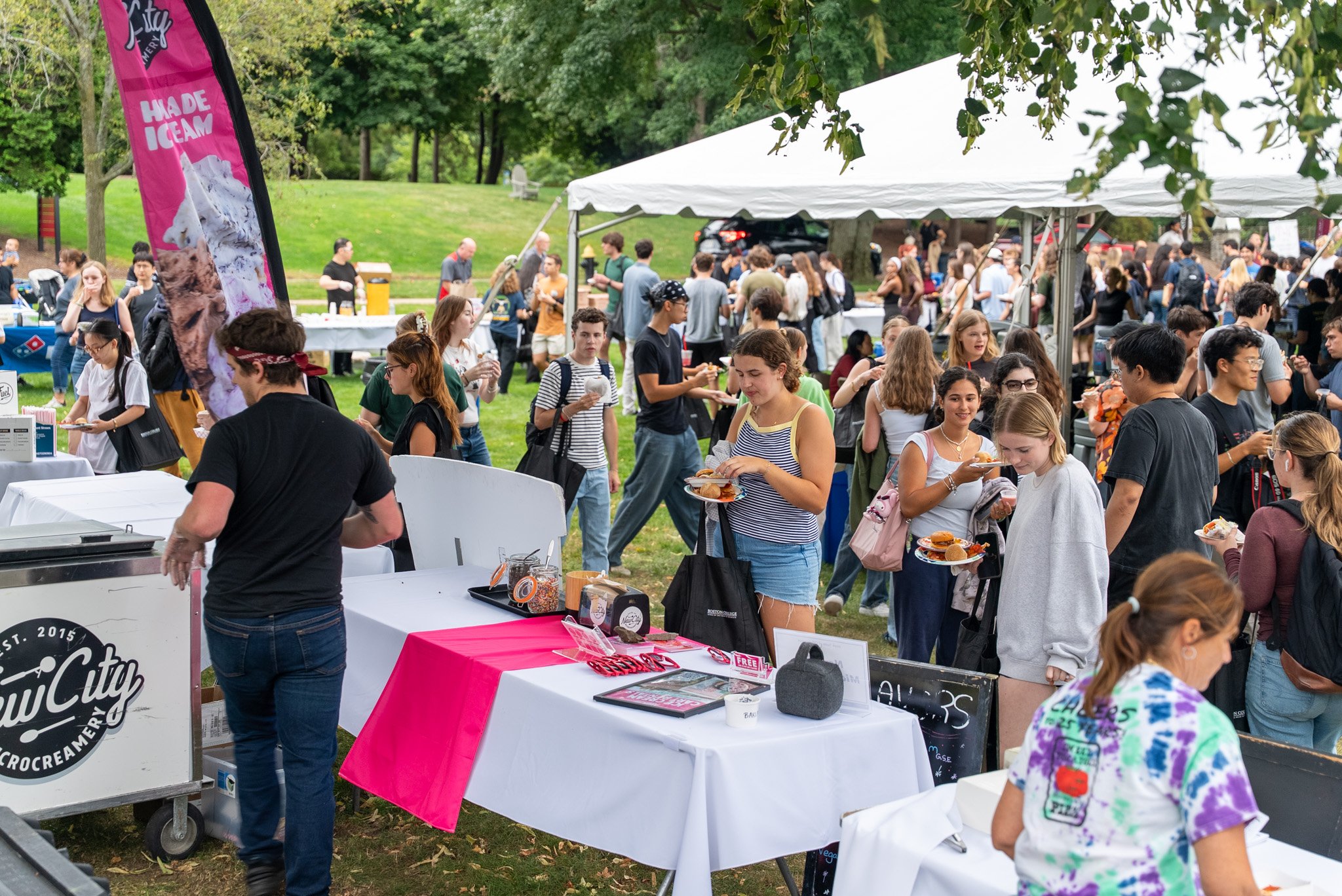 Crowd of people at an outdoor event with food stalls and tents in a park, surrounded by green trees.