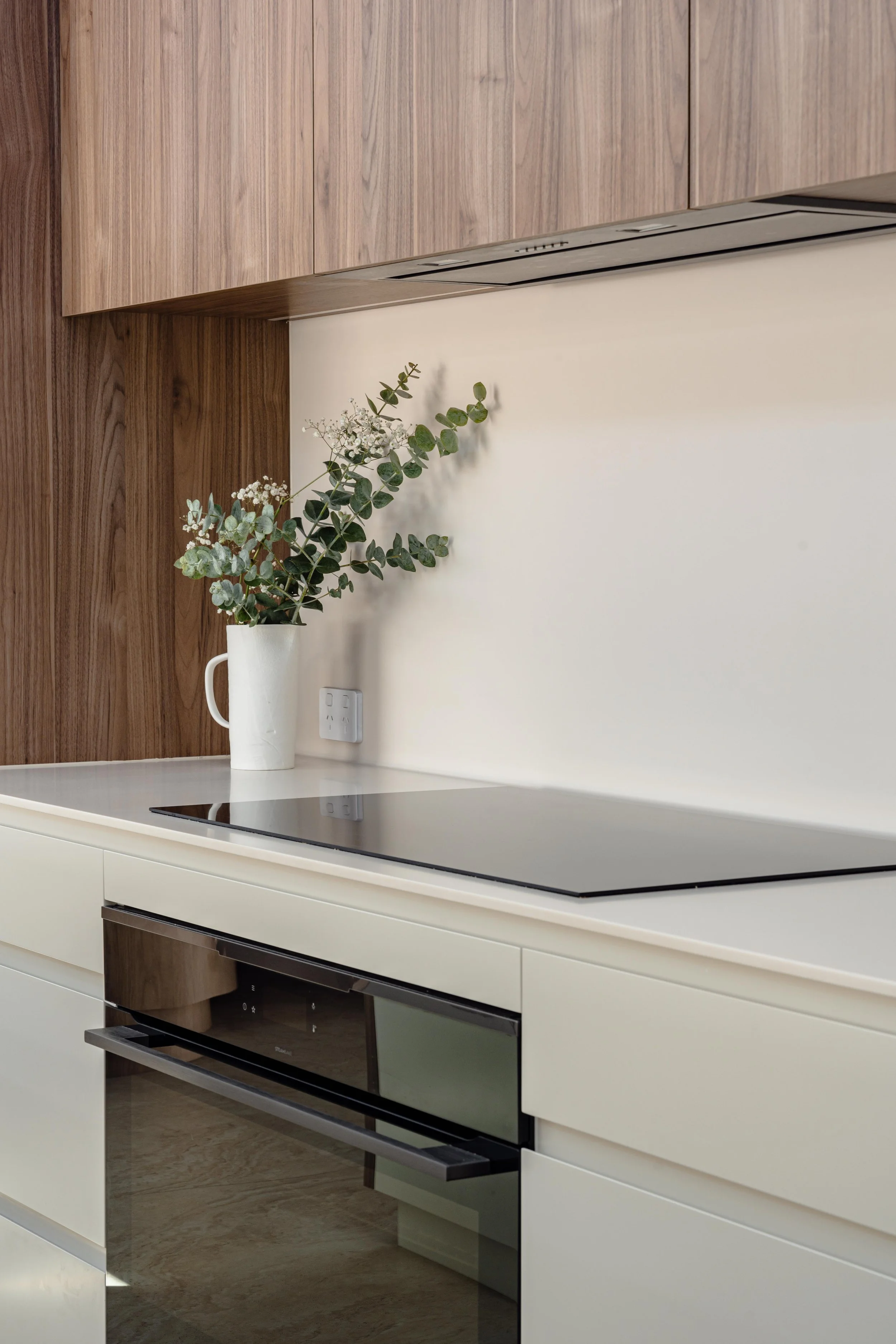Modern kitchen with white countertop, black stovetop, wooden cabinets, and a white vase with greenery.