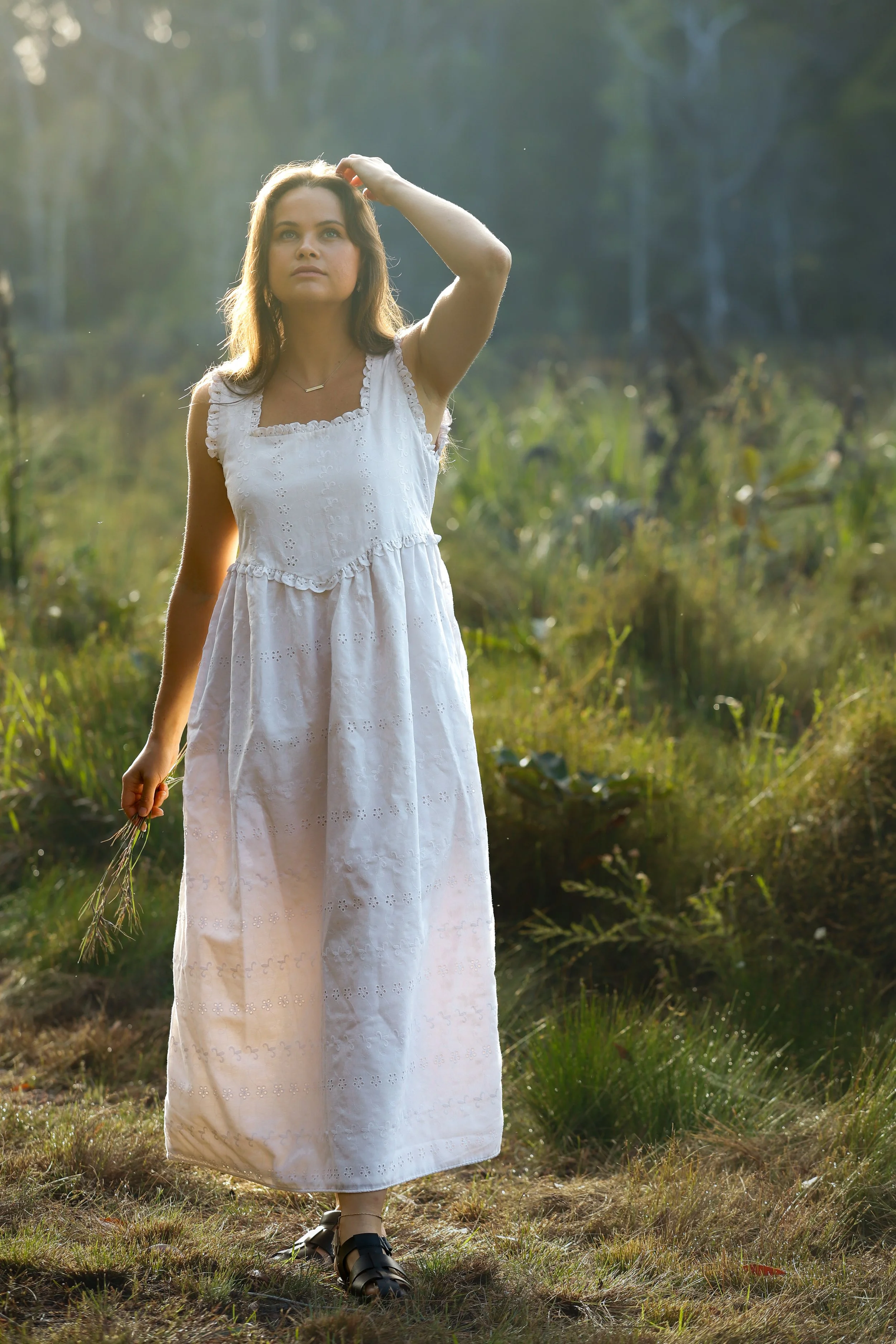 A woman dressed in a long white dress with eyelet embroidery walks outdoors in a grassy area, holding a bunch of grass in her left hand, and touches her head with her right hand. The background features blurred trees and sunlight filtering through the foliage.