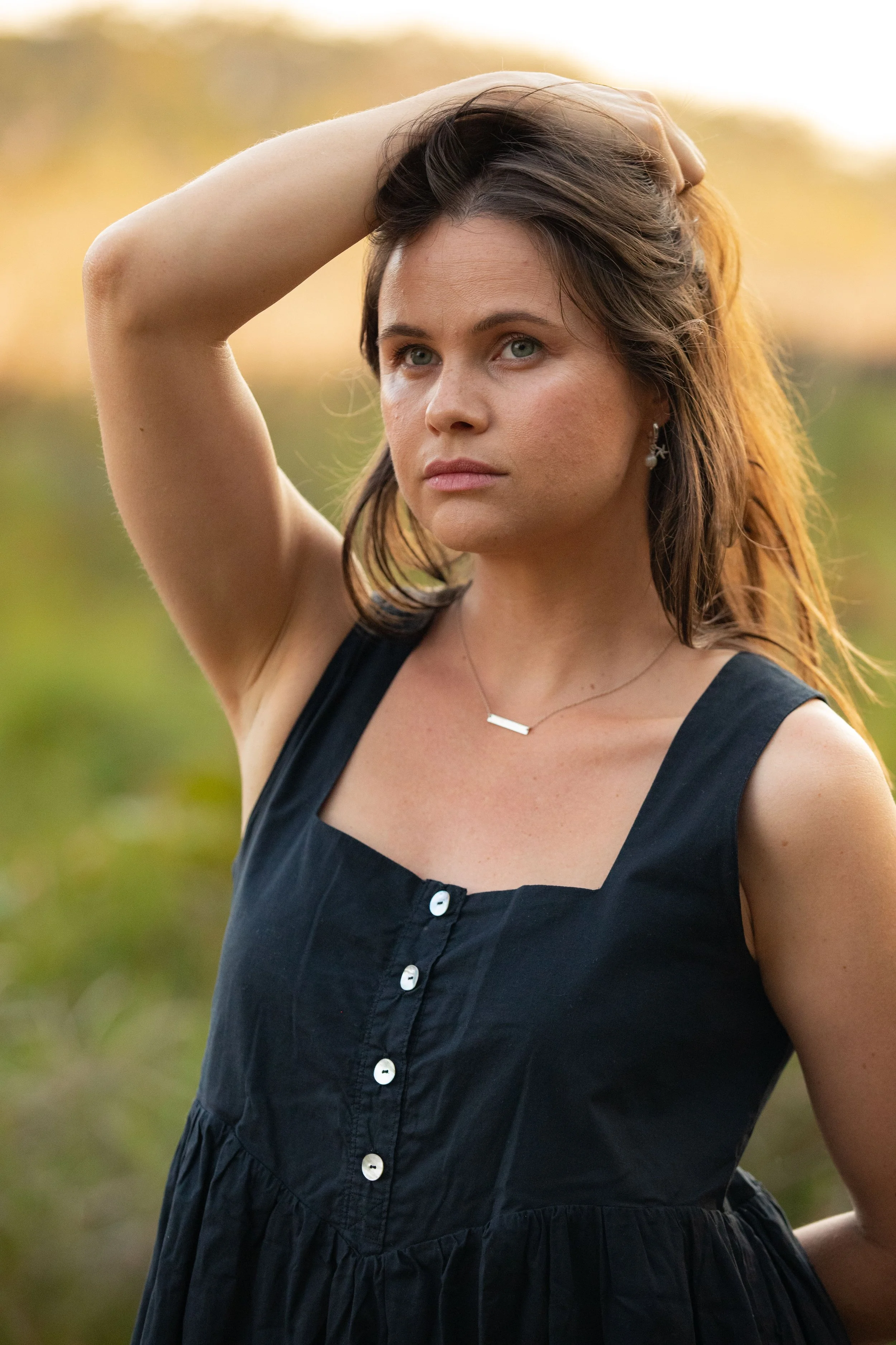 A woman with brown hair and blue eyes poses outdoors with her arm raised behind her head. She is wearing a black dress and silver earrings, with a blurred natural background.