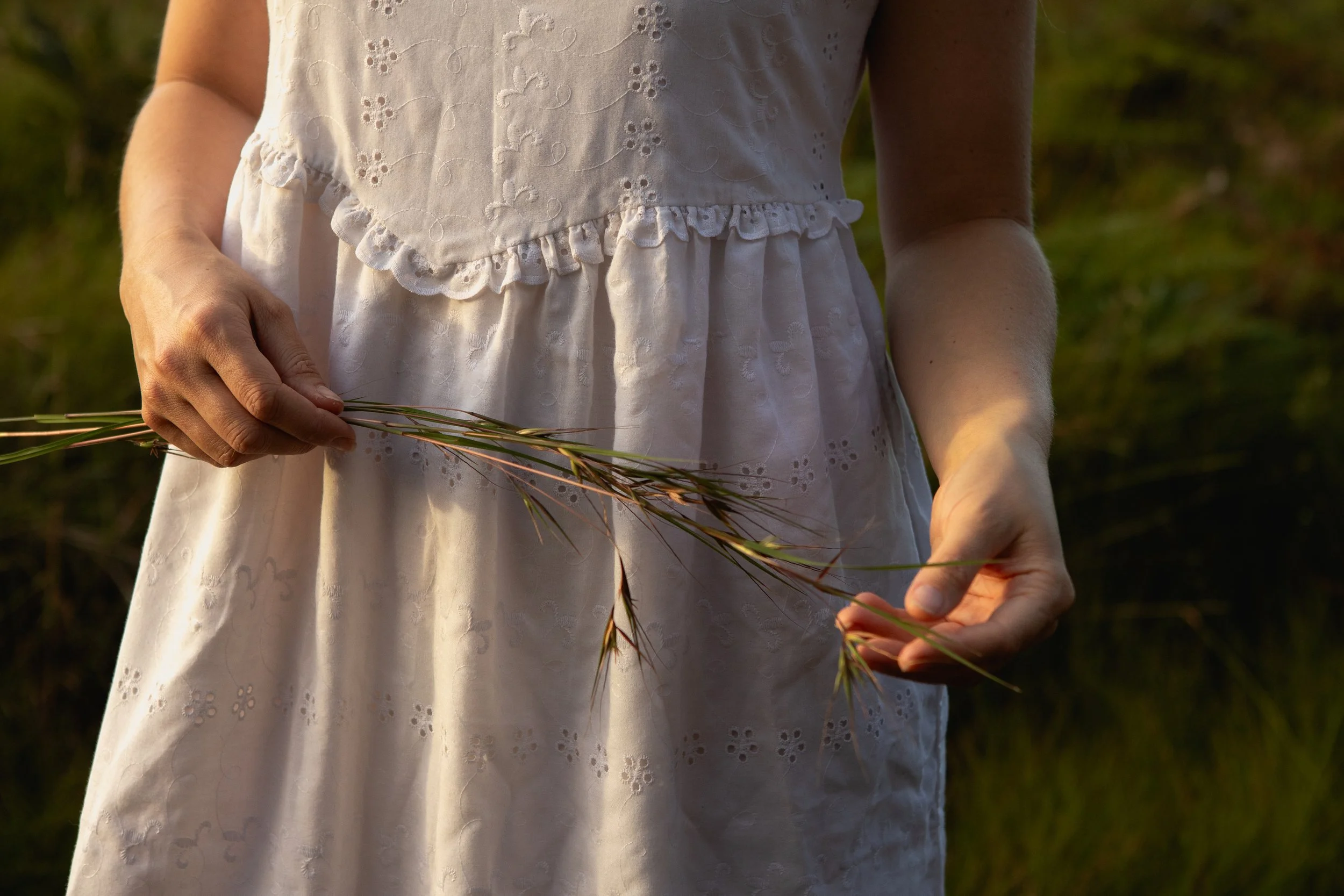A woman in a white dress with embroidered floral patterns is outdoors, holding a small bundle of grass or wheat.