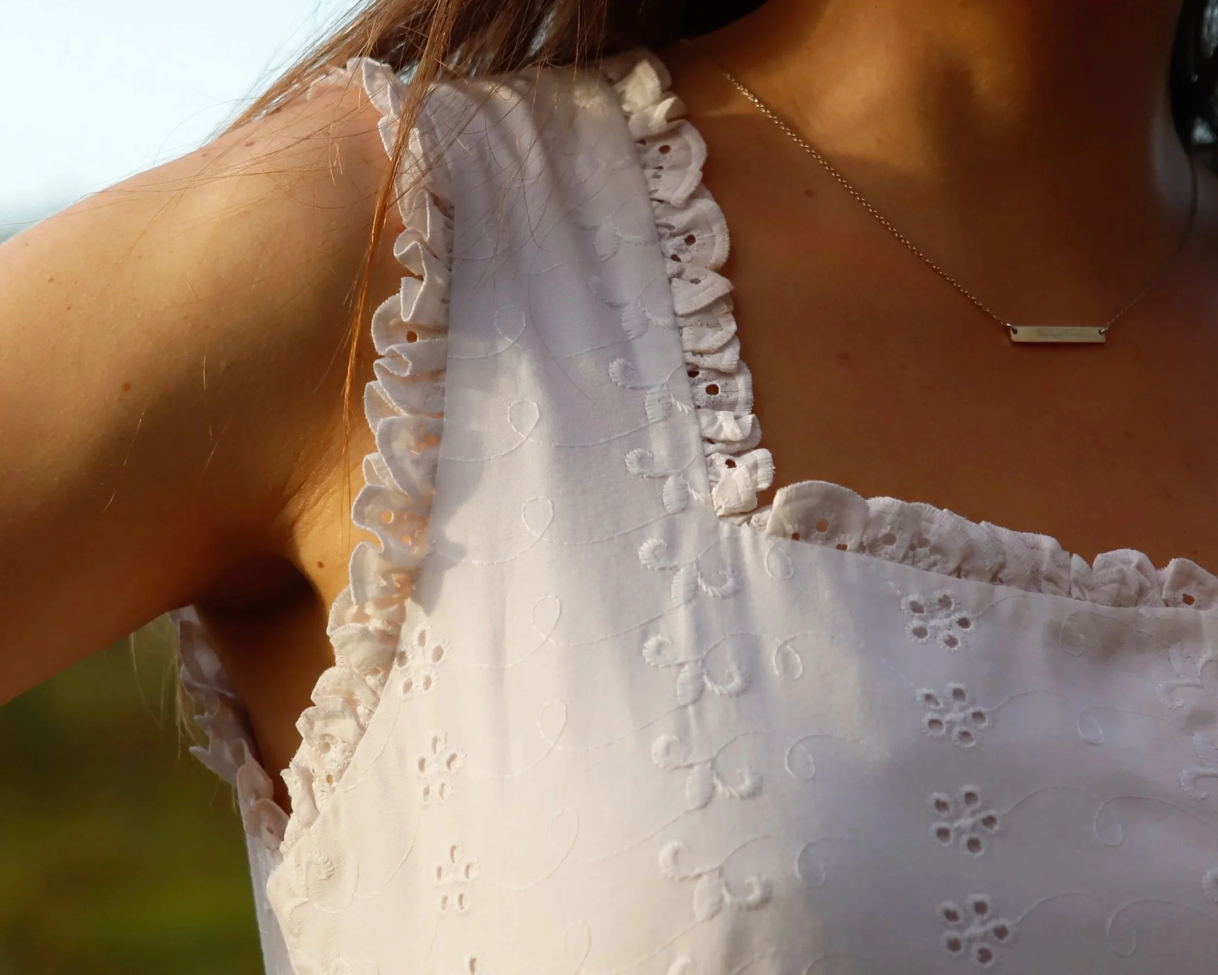 Close-up of a woman's neckline showing a delicate gold necklace with a rectangular pendant. She is wearing a white dress with eyelet embroidery and ruffled edges on the shoulders.