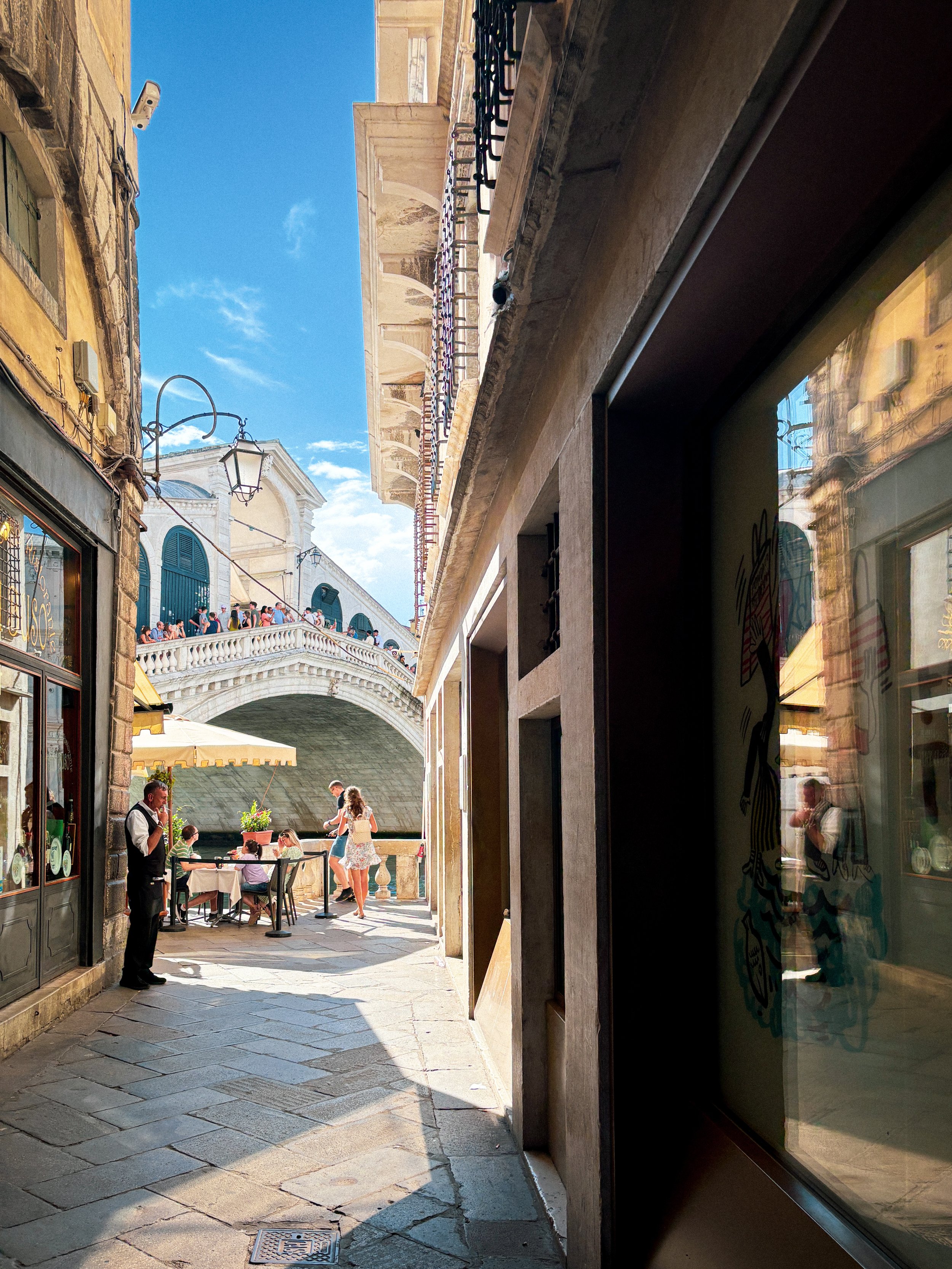 A narrow street scene in Venice, Italy, with a view of the Rialto Bridge over a canal, people dining outdoors at a restaurant, and buildings with balconies and shutters.