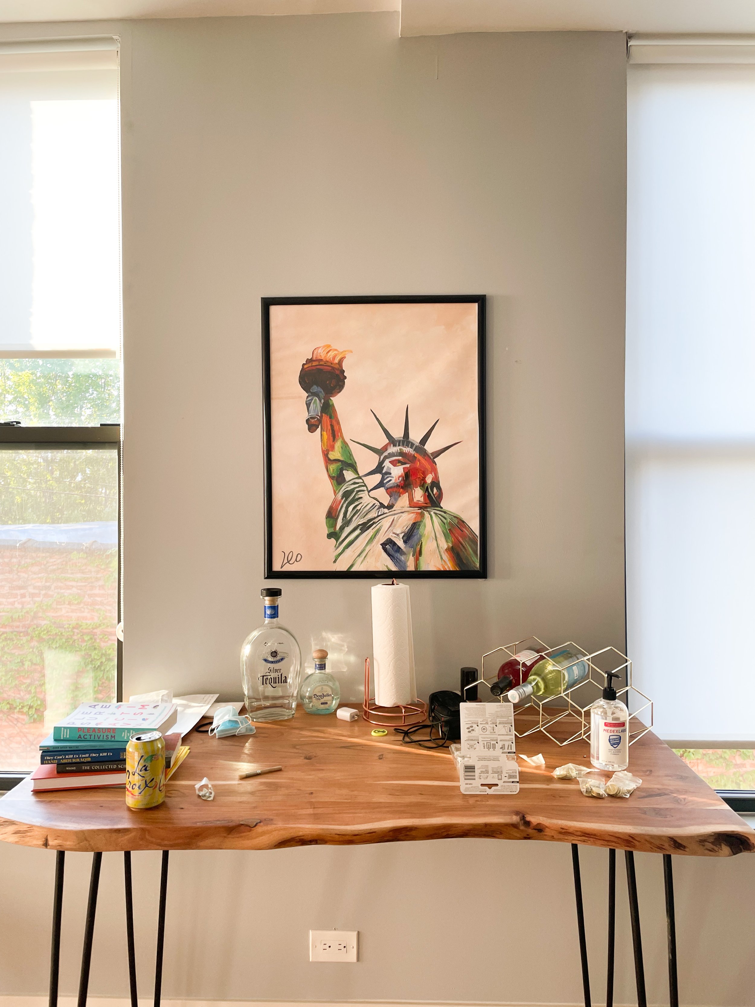 A wooden table with various items including books, a soda can, bottles of alcohol, a paper towel roll, hand sanitizer, a mask, a pen, and a small dish. Behind the table, there is a wall with a framed watercolor painting of the Statue of Liberty holdi