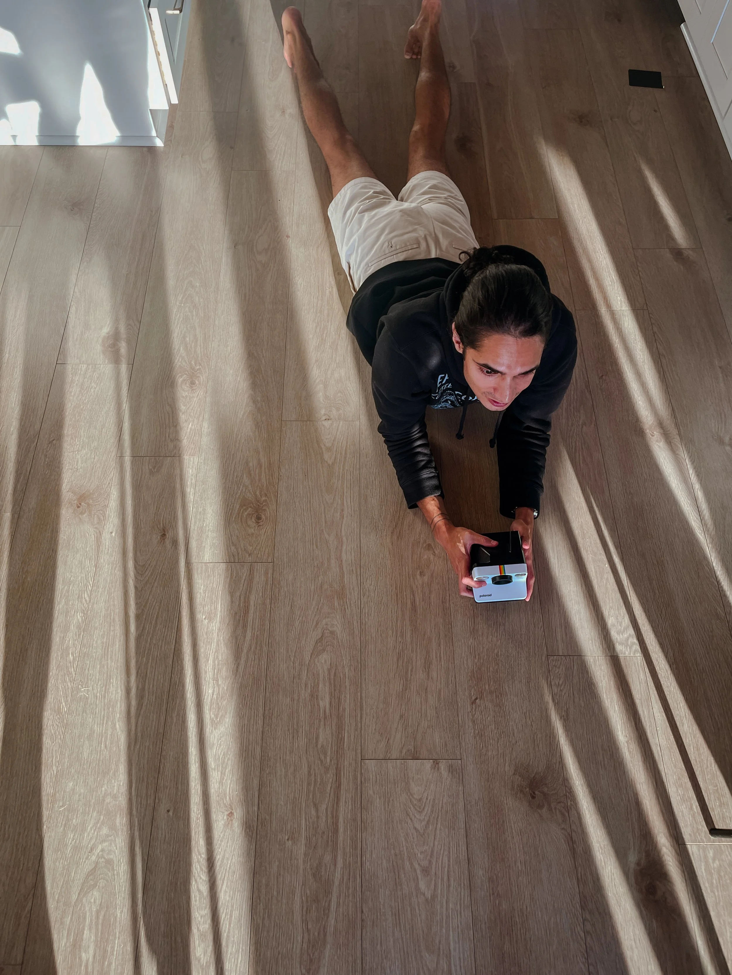 A woman lying on the wooden floor indoors, taking a selfie with a smartphone, with sunlight casting shadows across the floor.