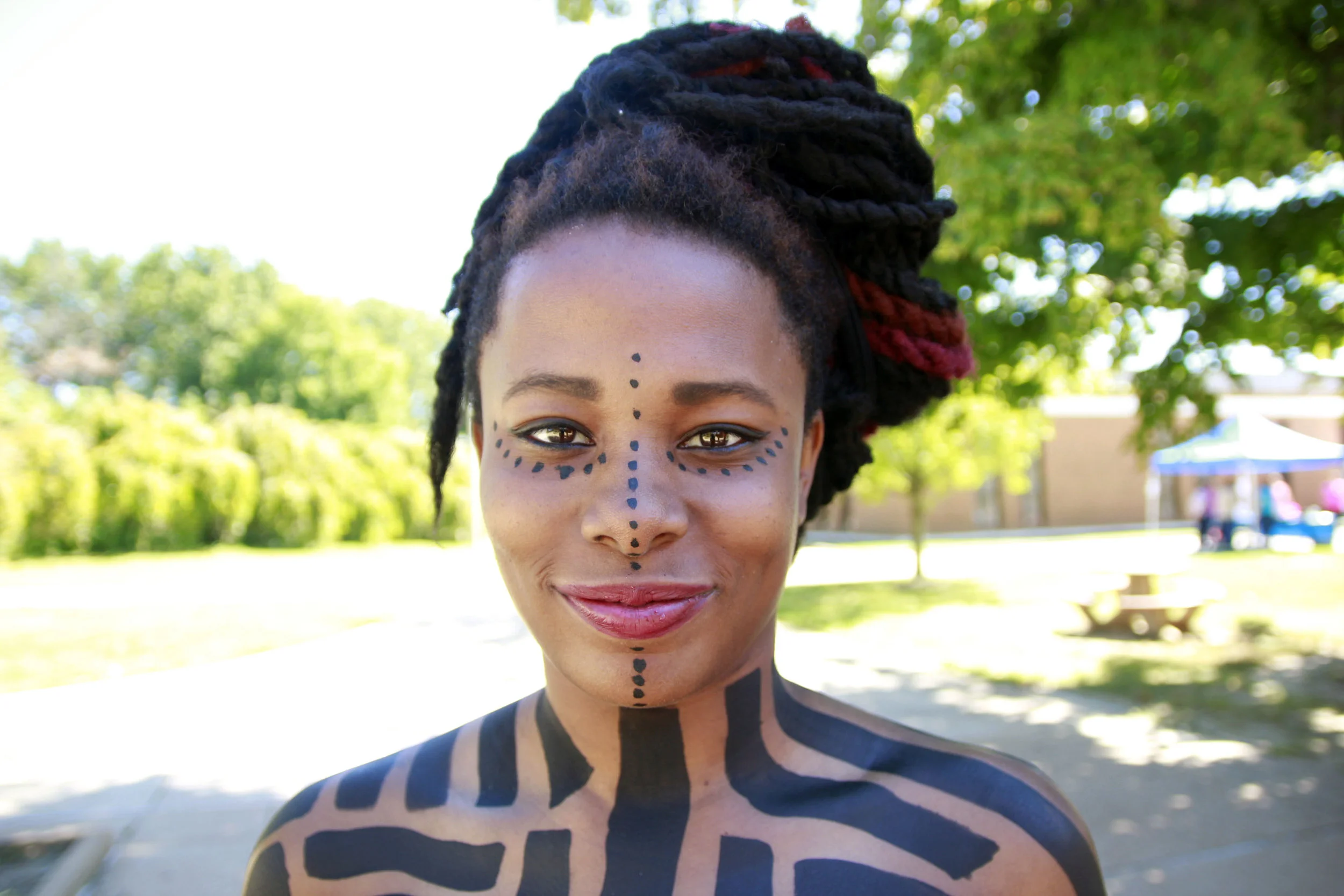 A woman with artistic face and body paint, dreadlocks hairstyle, standing outdoors in a park on a sunny day.
